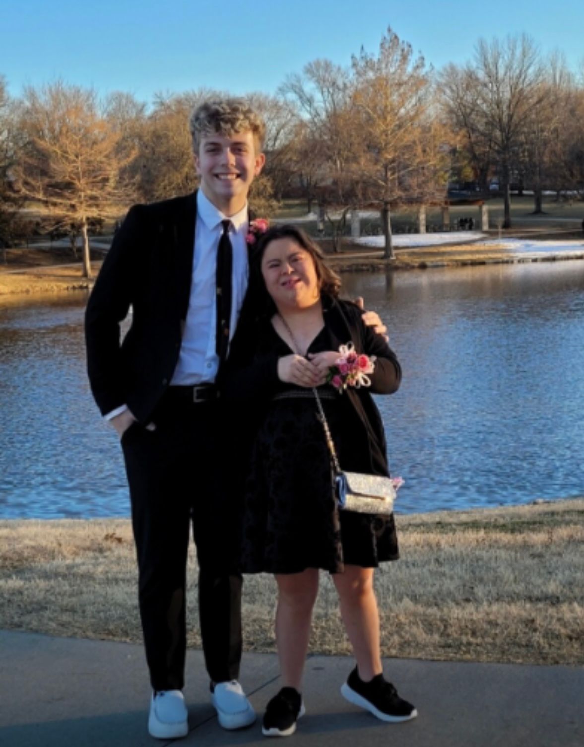 Two people in formal attire stand together by a scenic pond on a winter day.