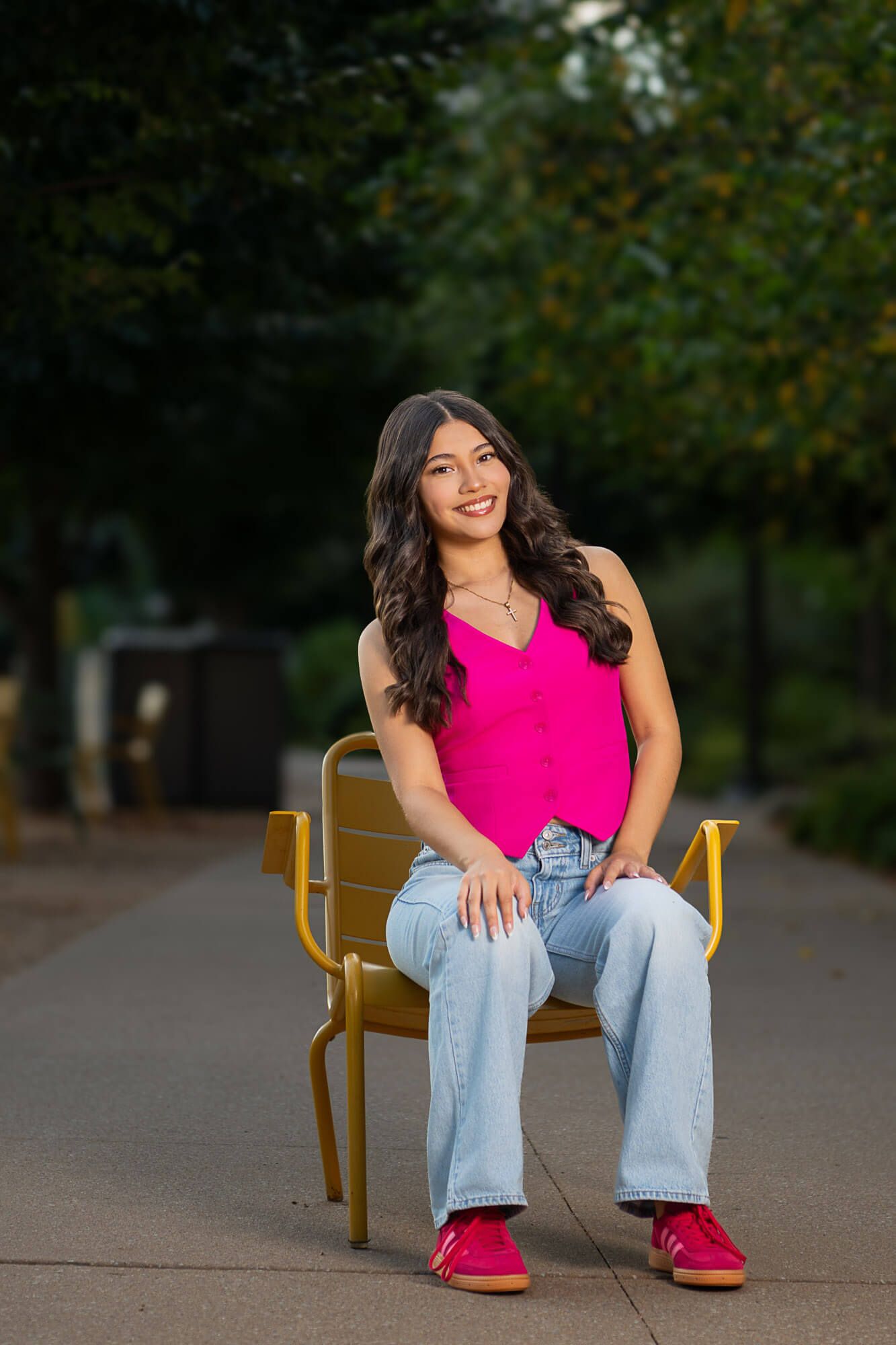 A casual outdoor portrait featuring a bright pink tank top, relaxed jeans and red shoes while sitting in a yellow chair.