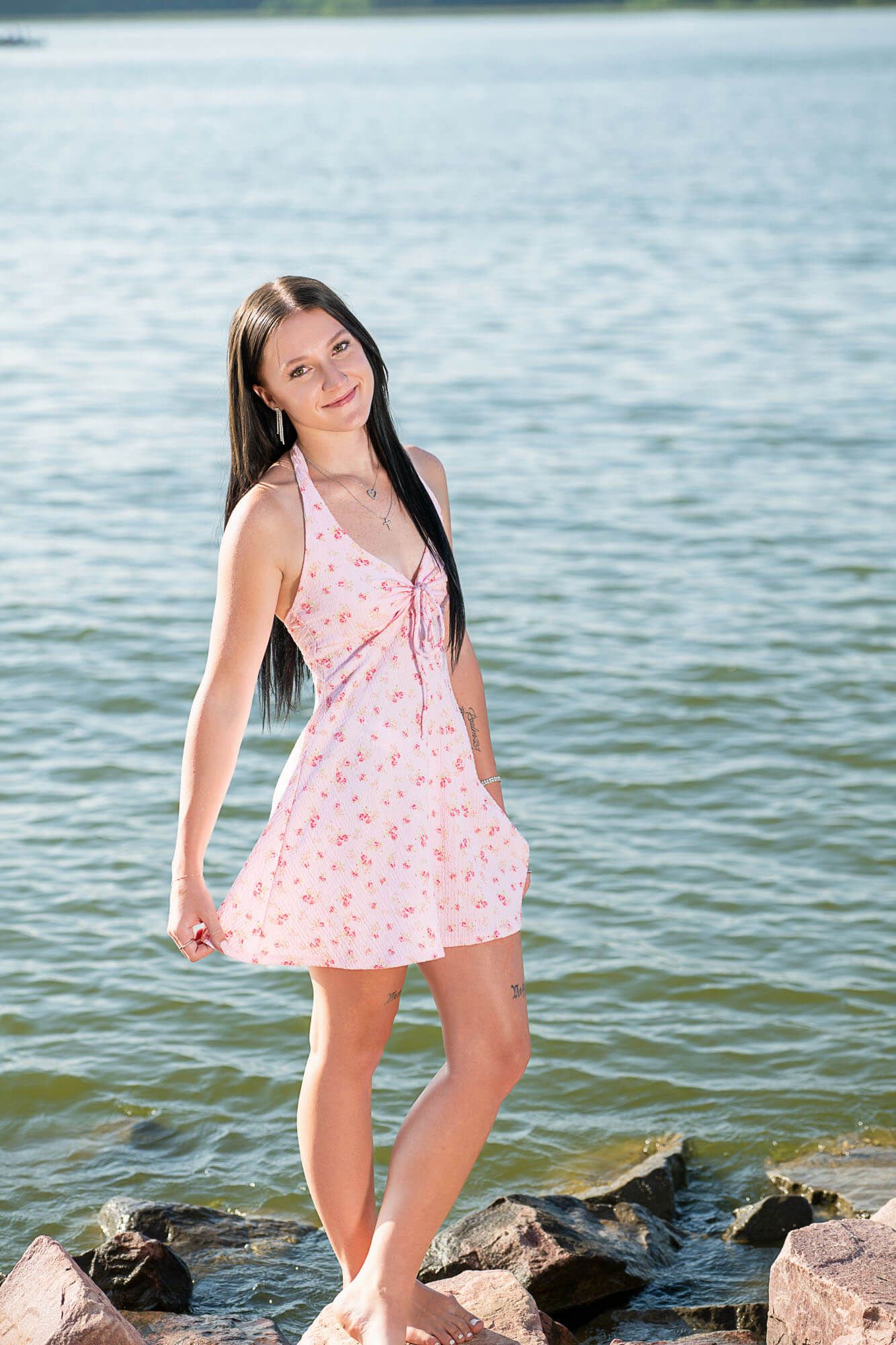 A young girl in a pink floral dress poses playfully near the water's edge on rocky shoreline.