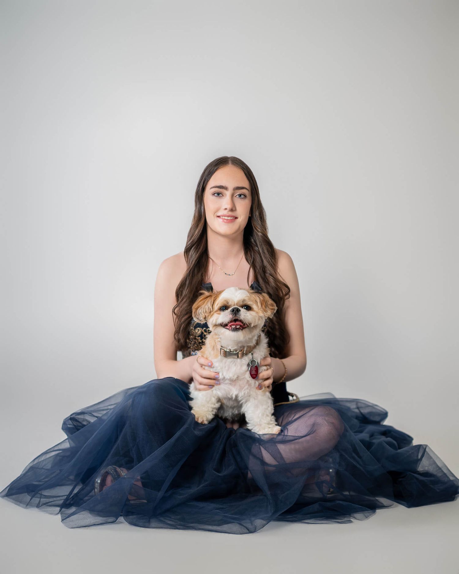 A person in a navy blue tulle gown sits gracefully with a small dog on a light gray studio backdrop.