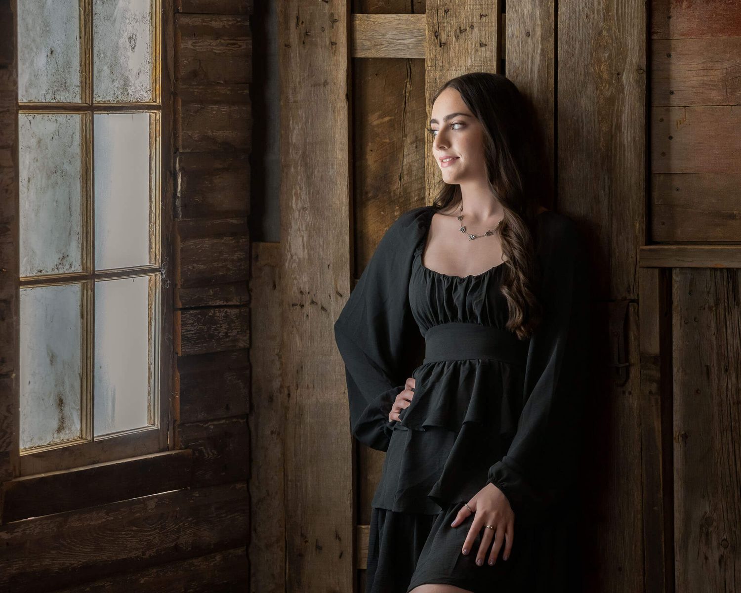 Omaha senior photography of female in a black dress poses in a rustic wooden room with natural window light creating a moody atmosphere.