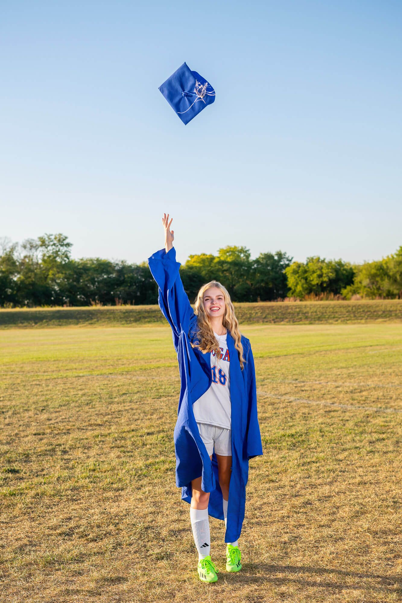 Smiling blonde graduate in blue cap and gown tossing her mortarboard in the air on a sunny green field.