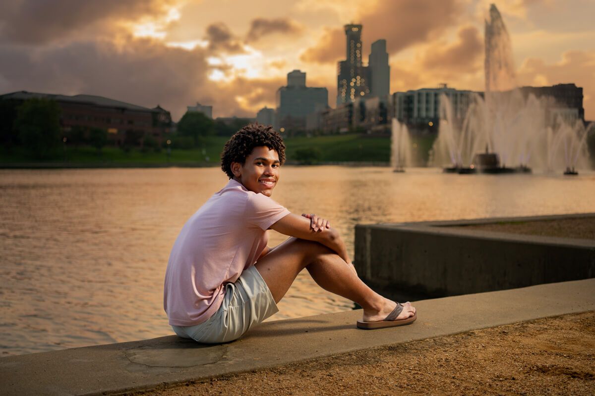 Young person sitting by a fountain at sunset with city skyline in the background.