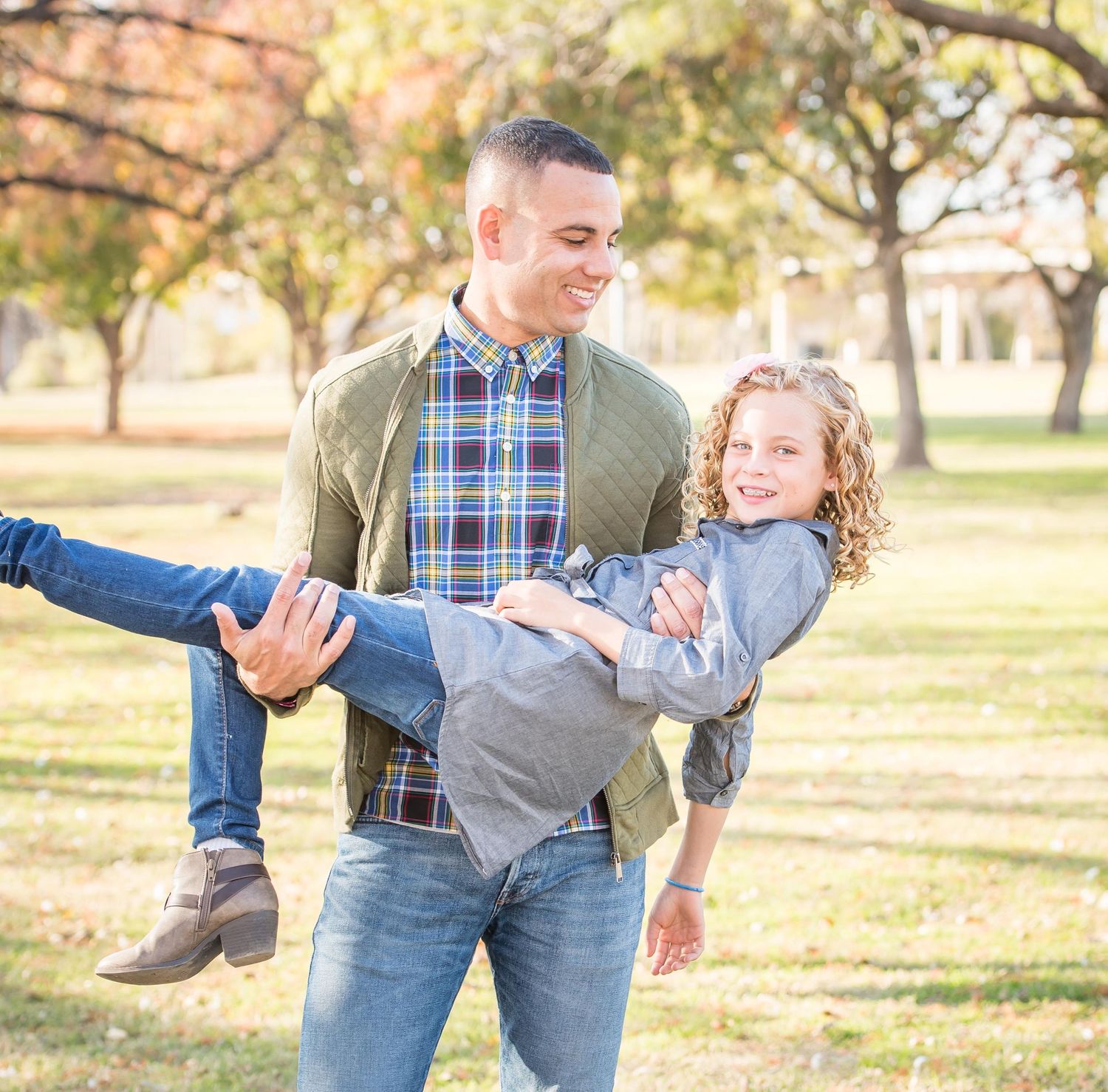 Dad and daughter enjoying playful moments in a rustic outdoor setting with autumn trees in the background by Omaha family photographer.