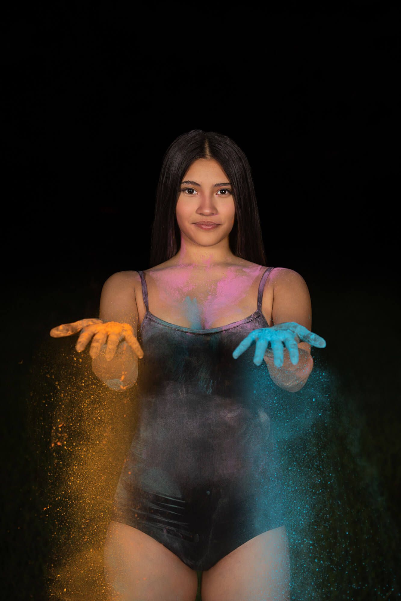 Young woman holding colorful Holi powder in orange and blue hands against a dark background.