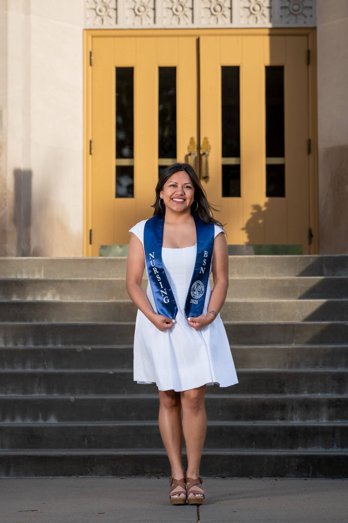 Smiling graduate in white dress and blue nursing sash stands on steps before yellow doors for graduation photo.