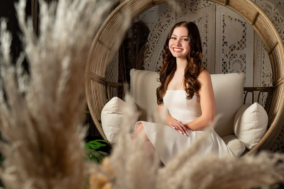 Young woman in white dress smiling in a rattan egg chair with pampas grass decor in a boho studio.