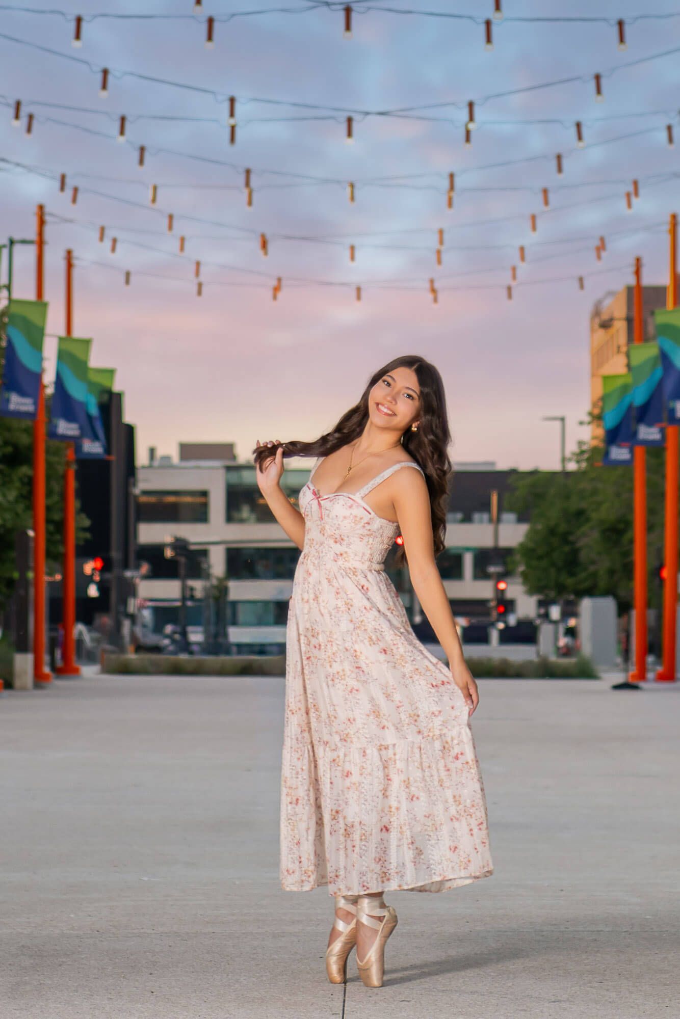 A graceful pose in a flowing floral dress beneath strung lights at dusk in an urban setting.
