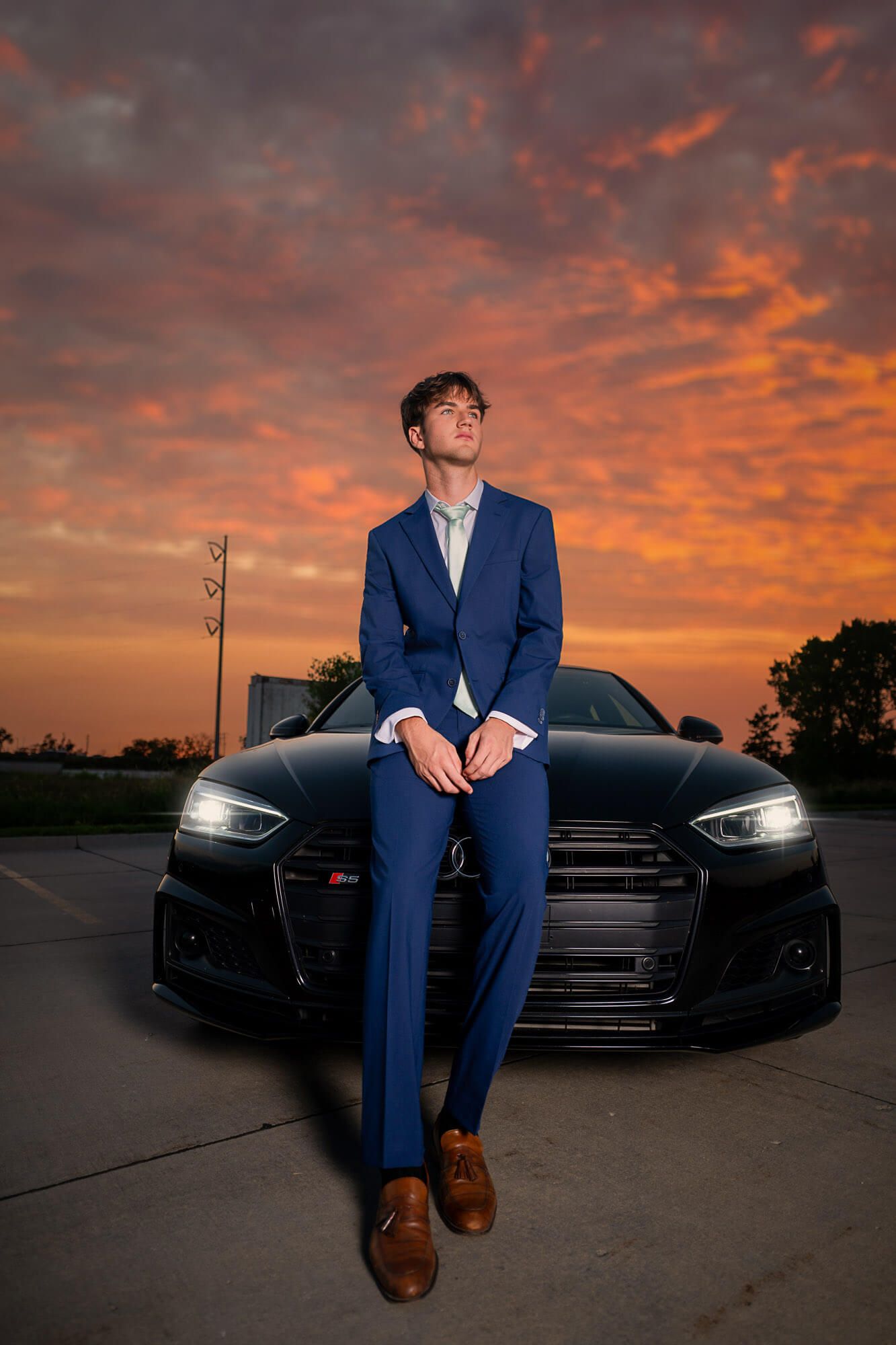 A person in a blue suit poses against a dramatic orange sunset sky while leaning on a black Audi car.