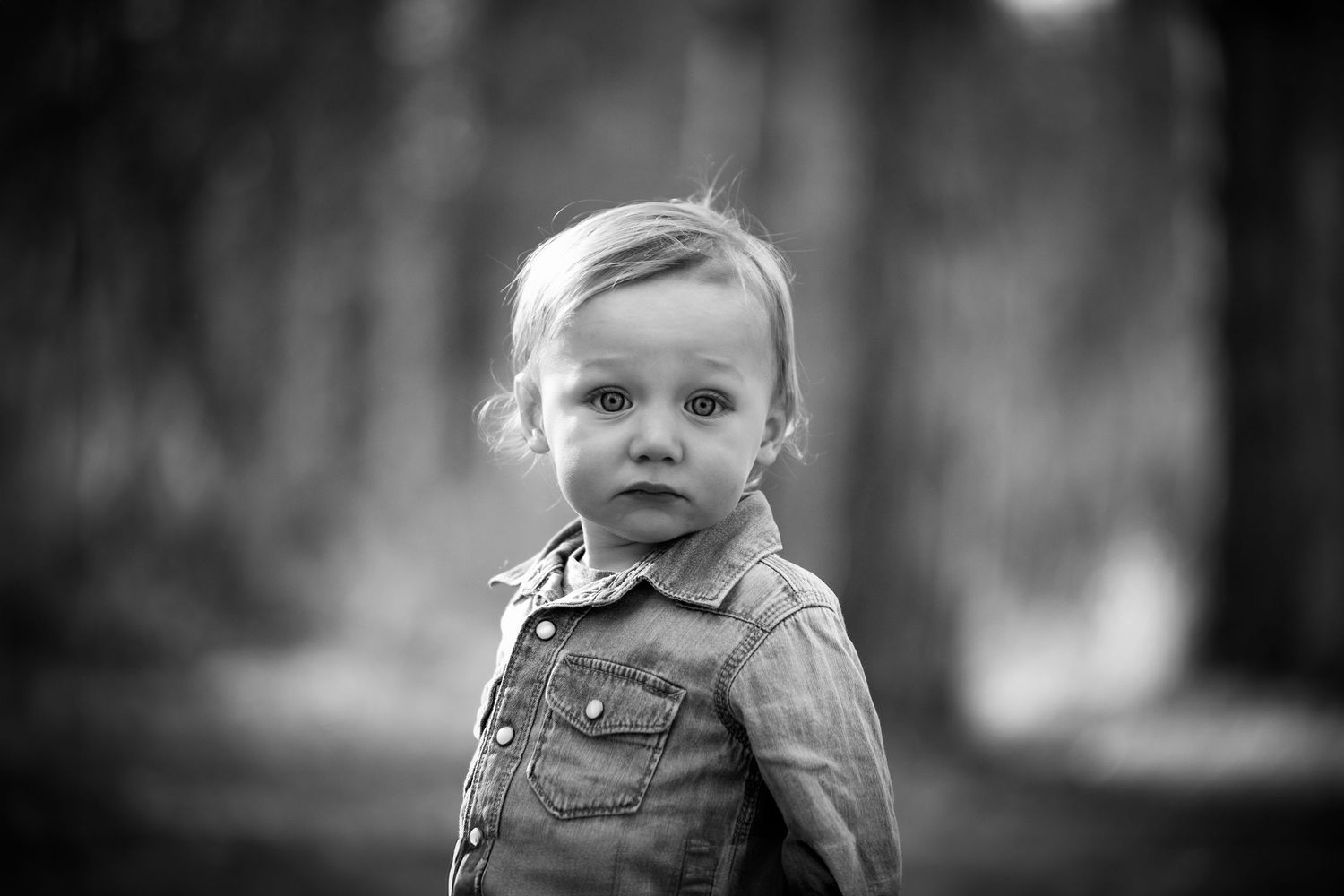 A toddler wearing denim overalls stands against a blurred monochrome background.