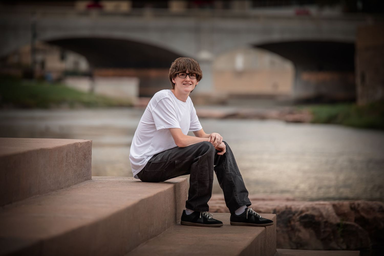 A person in a white t-shirt and dark pants sits on concrete steps near a bridge in soft evening light.
