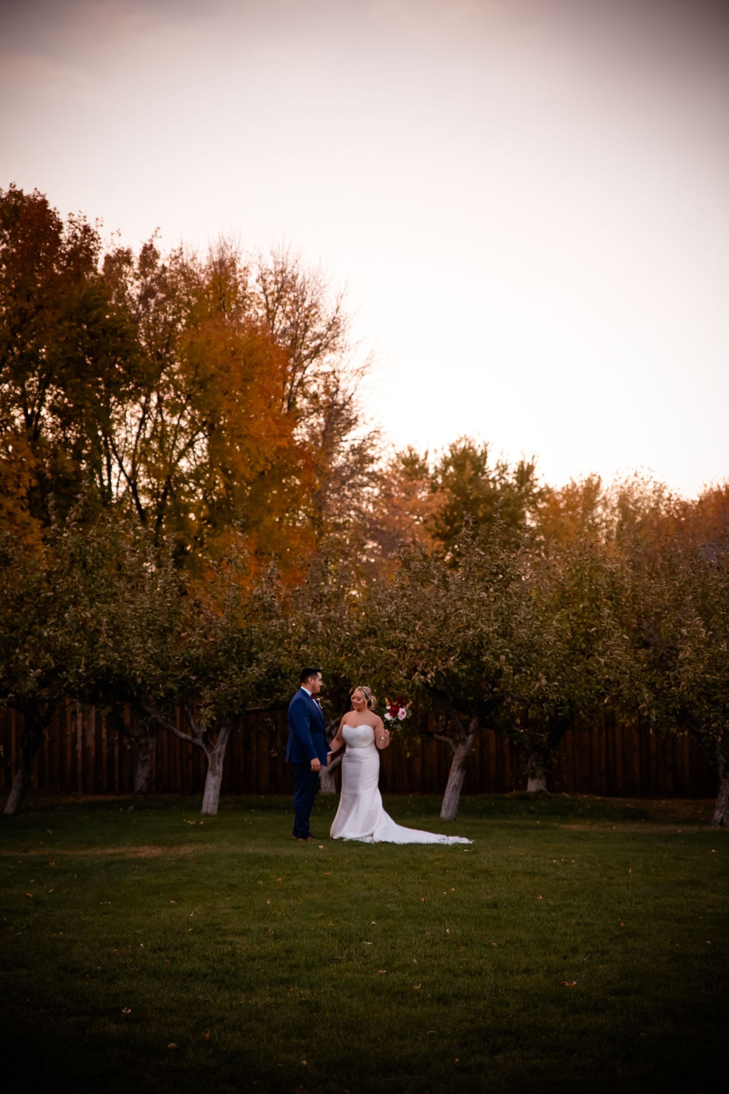 A couple stands together on a lawn surrounded by autumn trees during a sunset wedding ceremony.