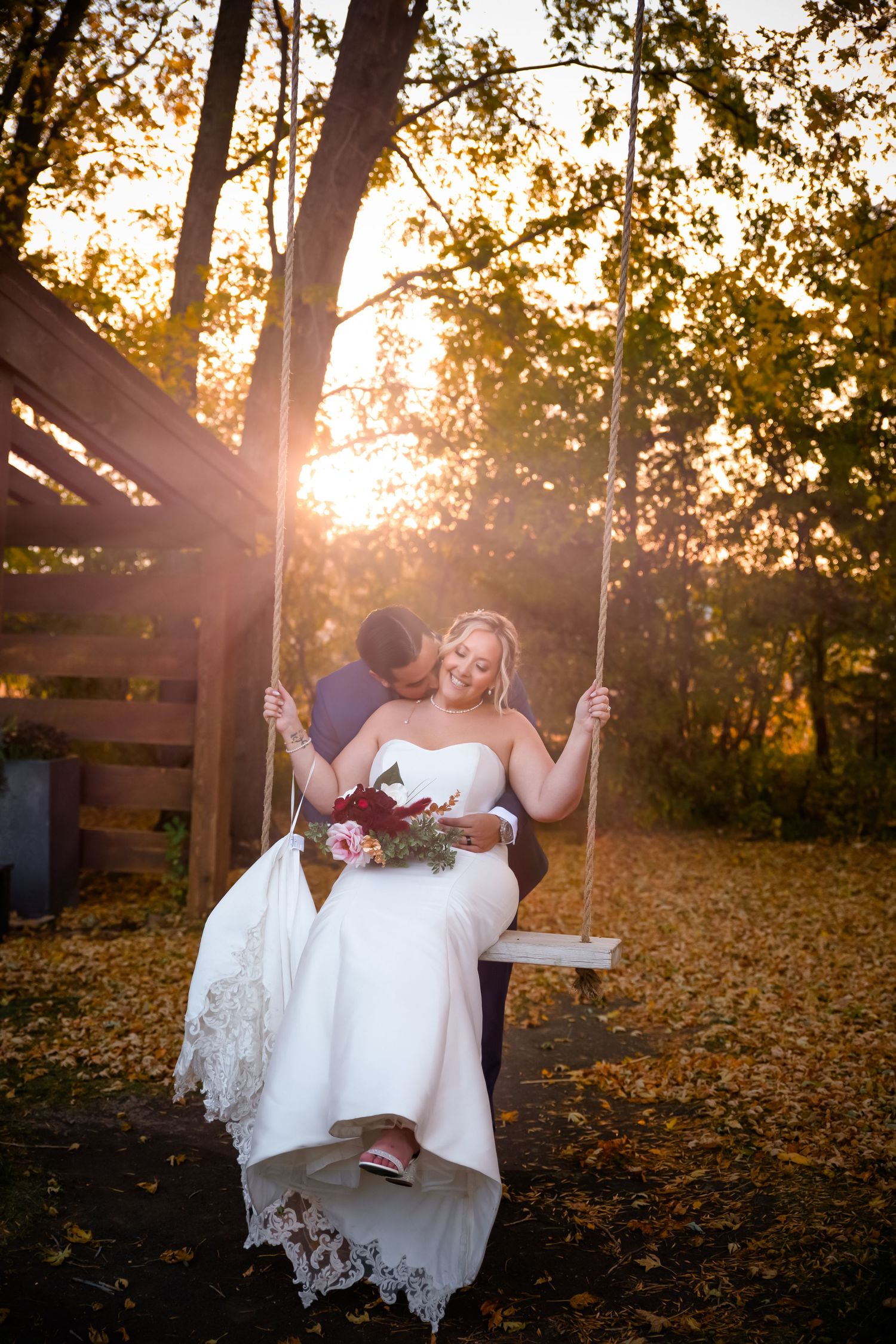 A couple on a wooden swing share a romantic moment at sunset with autumn leaves scattered on the ground.