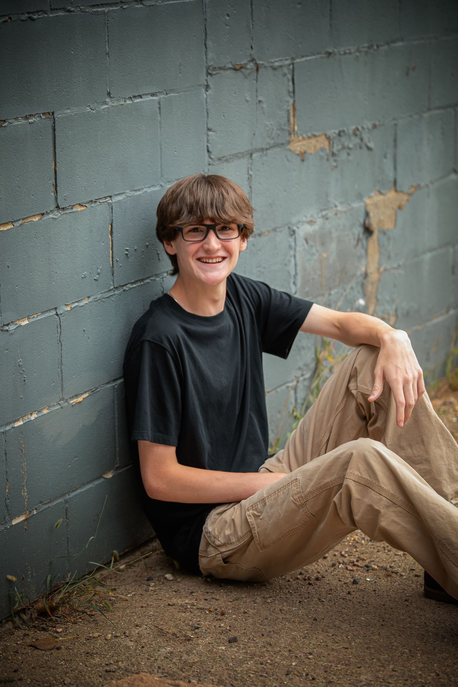 A series of casual portrait shots showing someone in a black t-shirt reclining against a gray wall and dirt ground.