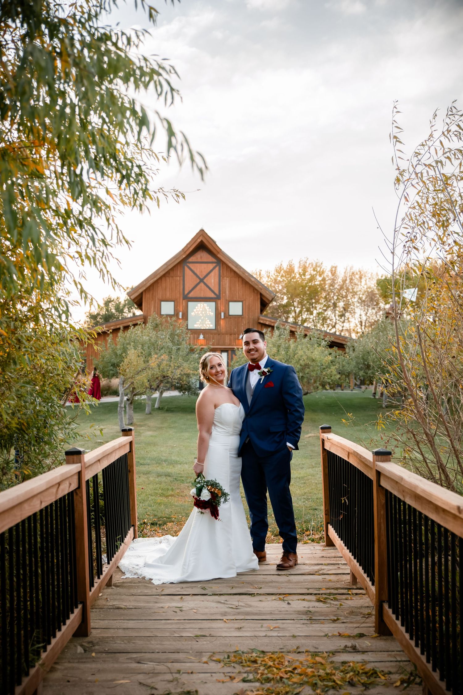 A couple stands on a wooden bridge with a rustic barn and autumn trees in the background during a fall wedding.
