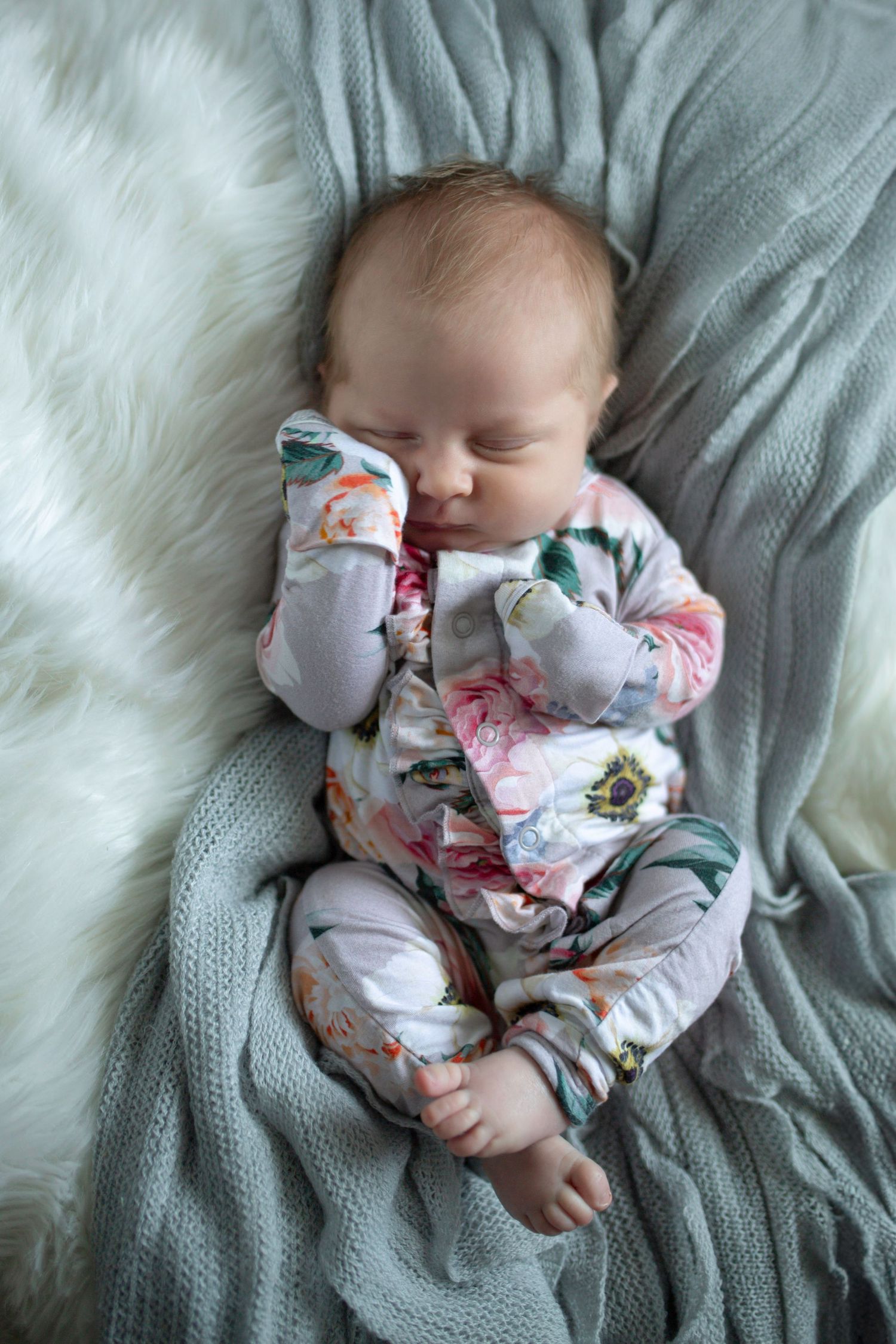 Sleeping baby in floral outfit lies on gray knit blanket.
