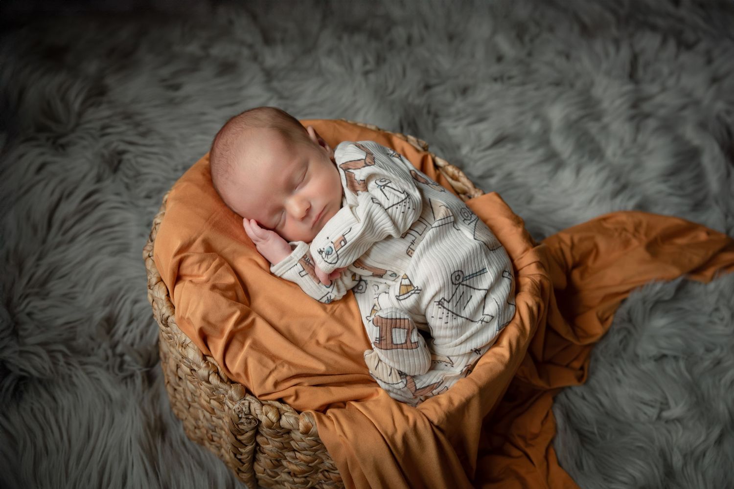 Sleeping newborn baby wrapped in newspaper print swaddle rests in tan basket on gray fur backdrop.