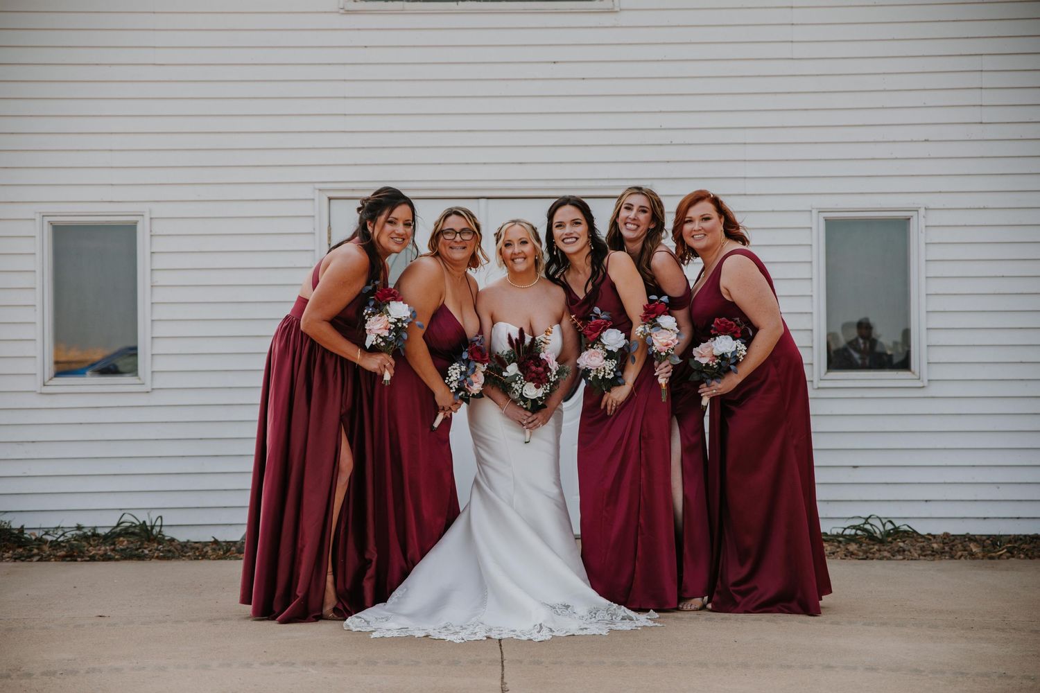 Bridal party in deep red dresses gathered for photos outside a white church.