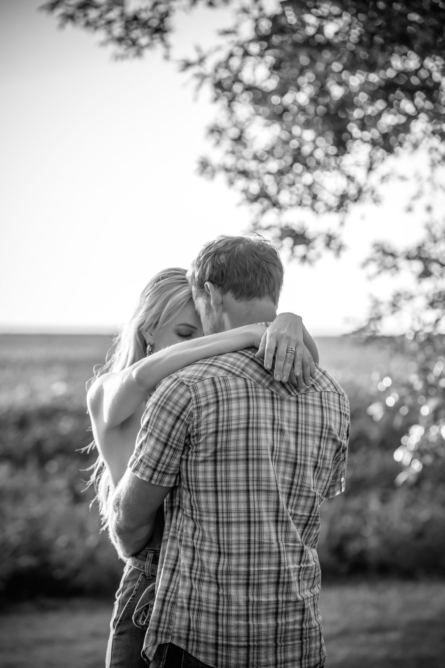 A romantic black and white photo of a couple embracing in nature with sunlit trees in the background.