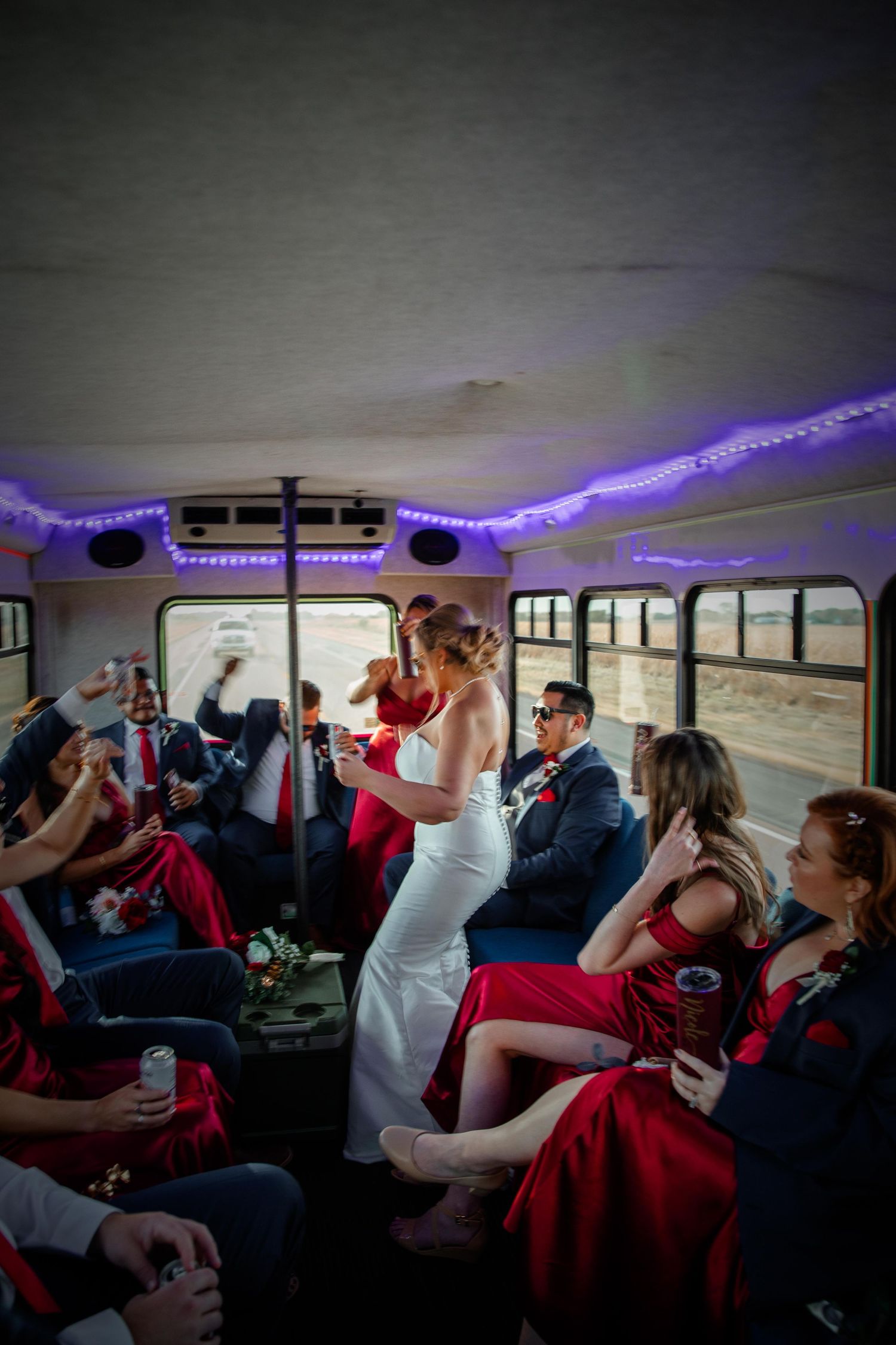 Wedding party celebrates inside a school bus decorated with purple LED lights during transportation.