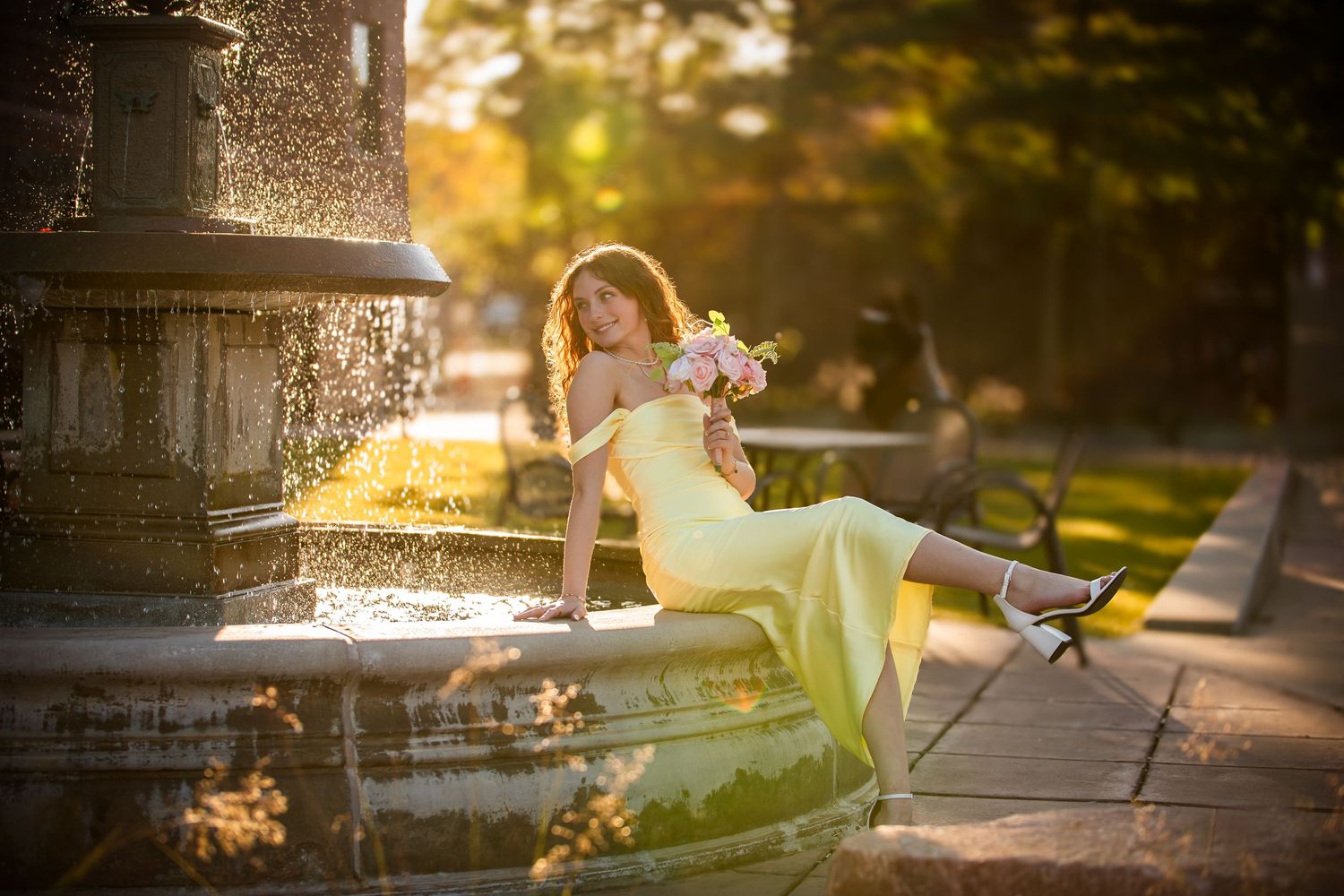 A person in a yellow dress sits on a fountain edge at sunset while water sprays in a golden-lit park setting.