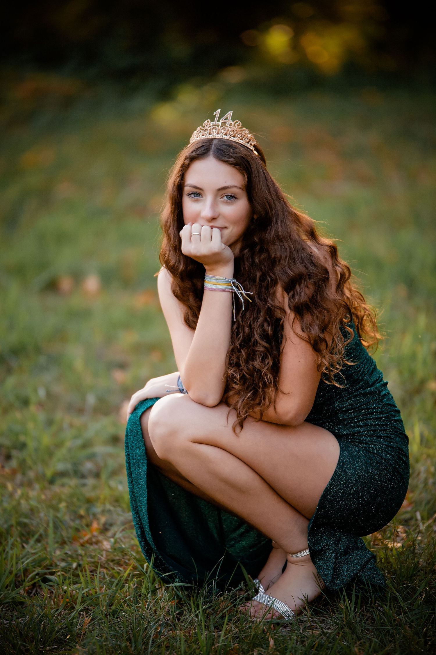 Young woman in green dress and tiara crouches on grass in outdoor setting.  Photographer in Sioux Falls, SD