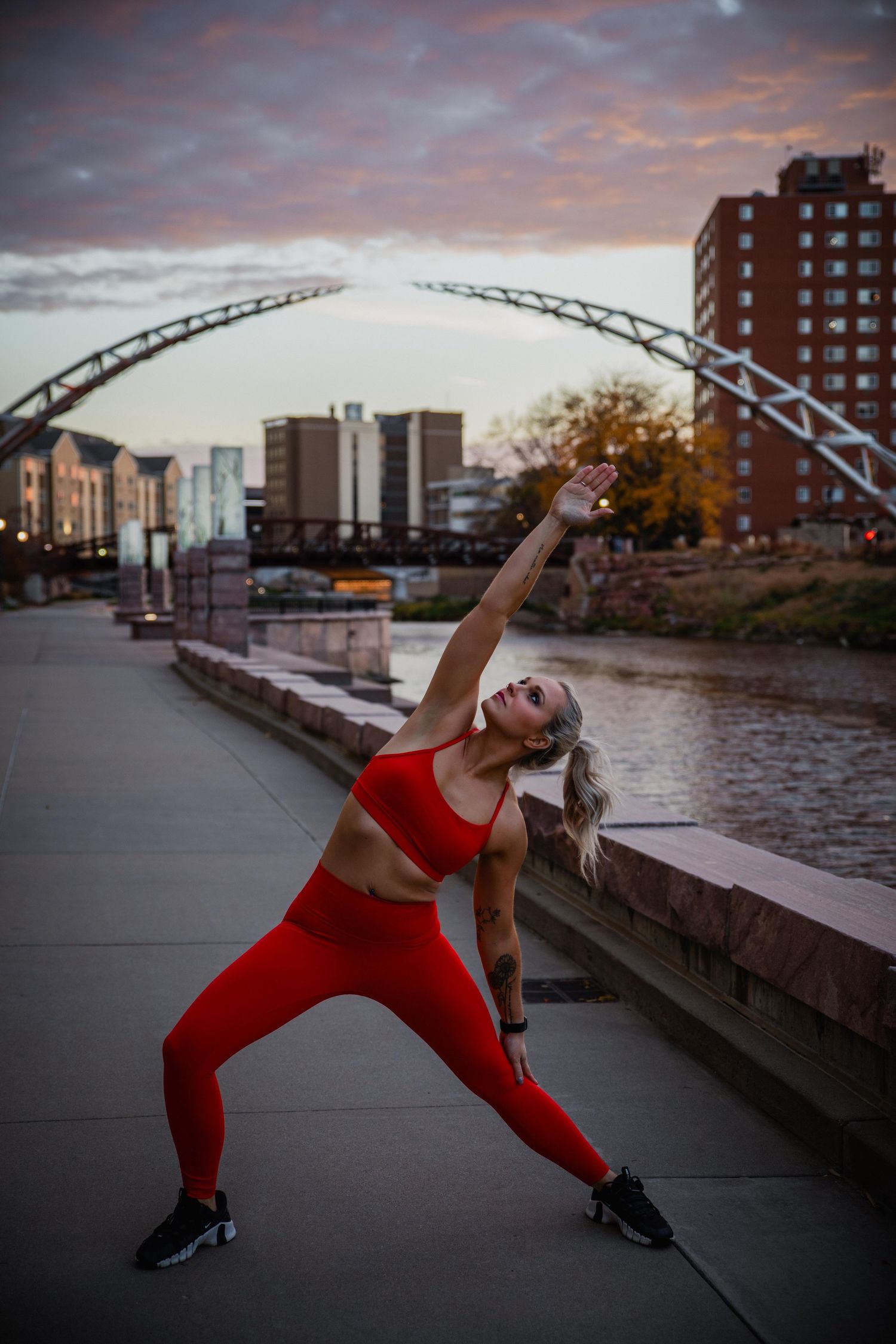 Dancer in red athletic wear strikes dramatic pose along riverside path at sunset with city bridge and buildings in background.
