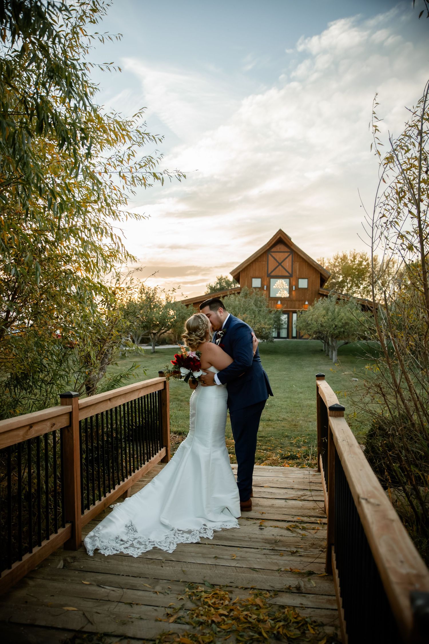 A romantic couple shares a tender moment on a wooden bridge during sunset at a rustic wedding venue.