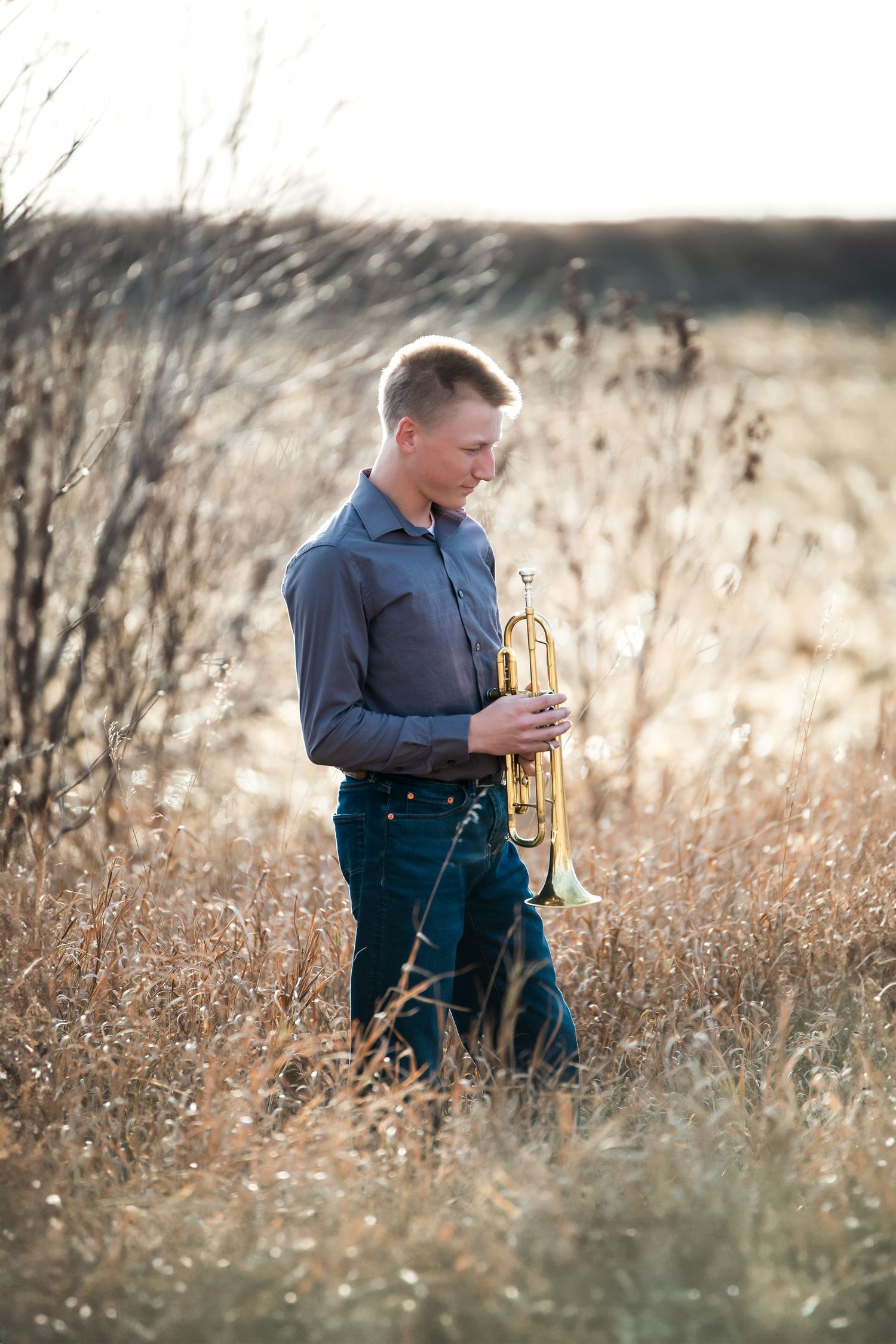 Young musician plays brass trumpet while sitting in tall autumn grass at sunset.