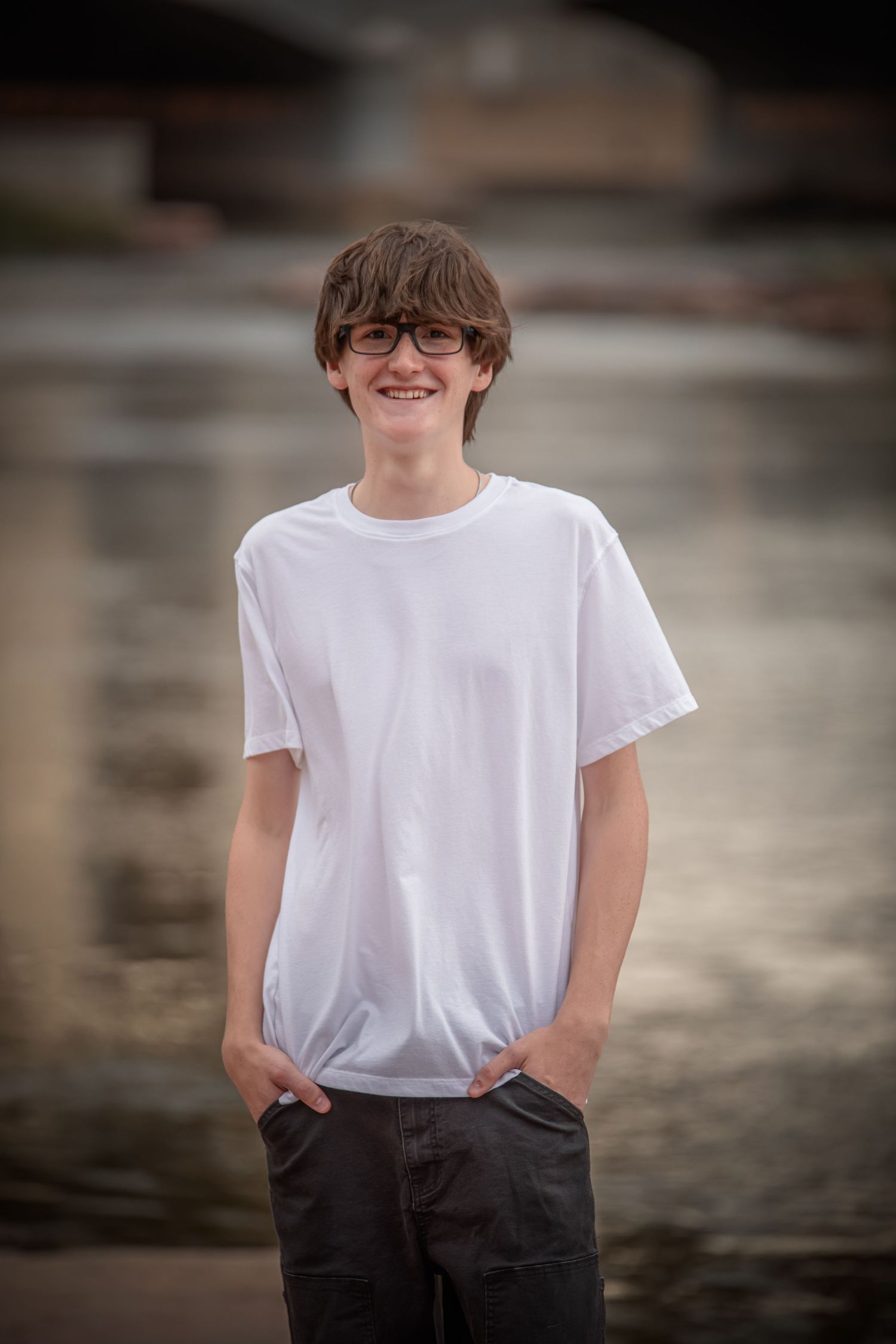 A casual portrait of someone wearing a white t-shirt, glasses and dark pants standing with hands in pockets.
