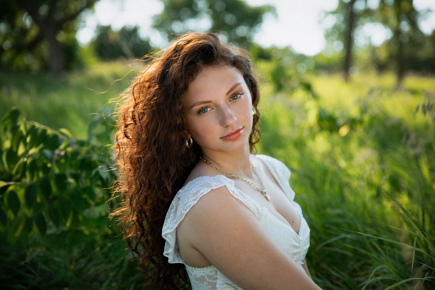 Young woman with curly auburn hair in white dress sitting in lush green field with sunlight filtering through trees.