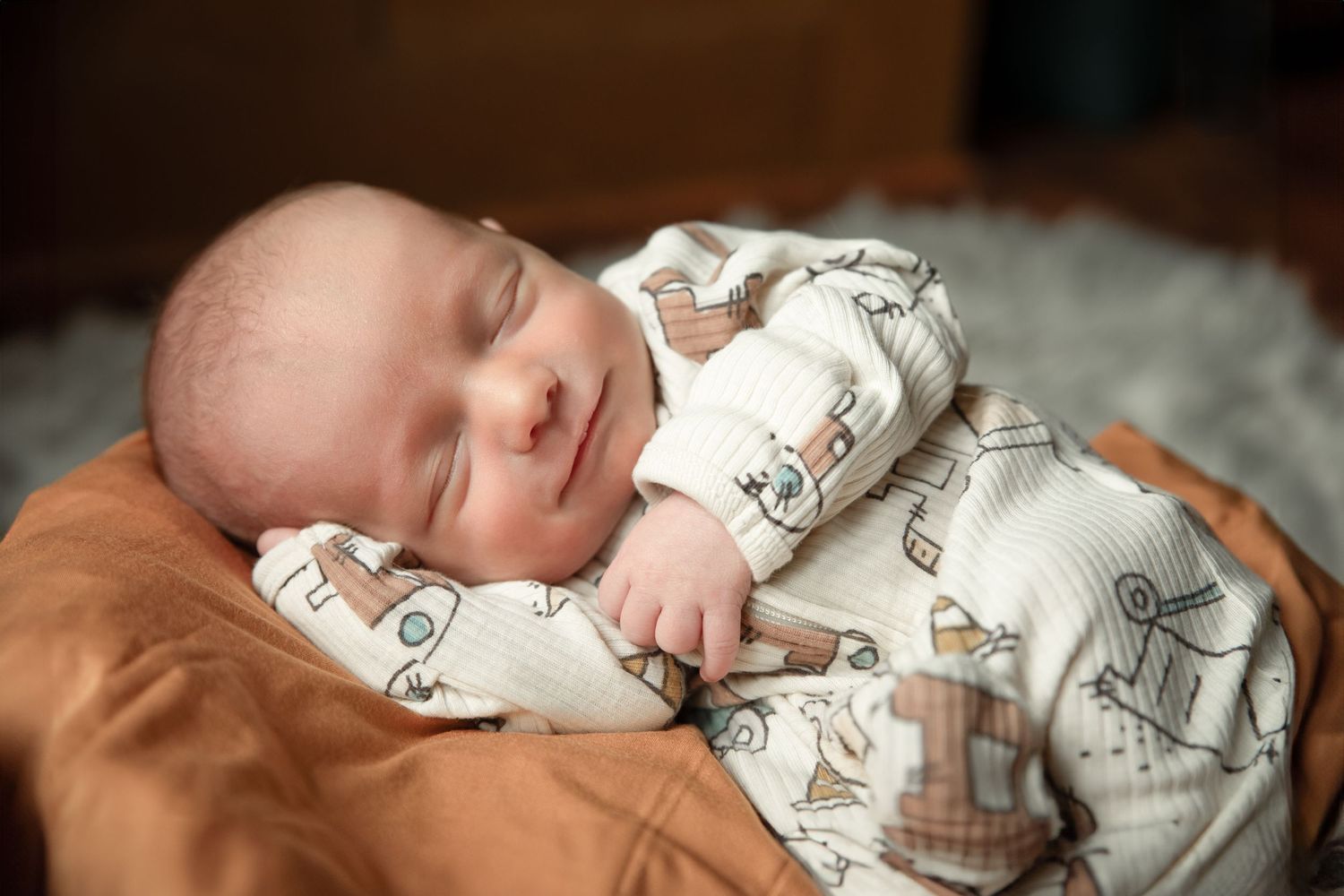 Sleeping baby wearing white patterned onesie rests peacefully on soft brown surface.