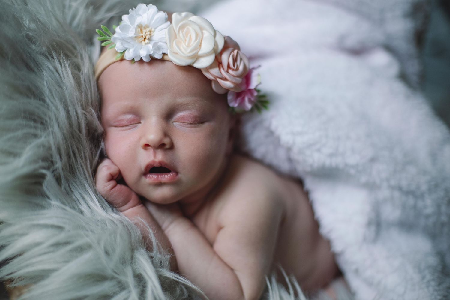 Newborn wearing floral headpiece sleeps peacefully on white fluffy backdrop.