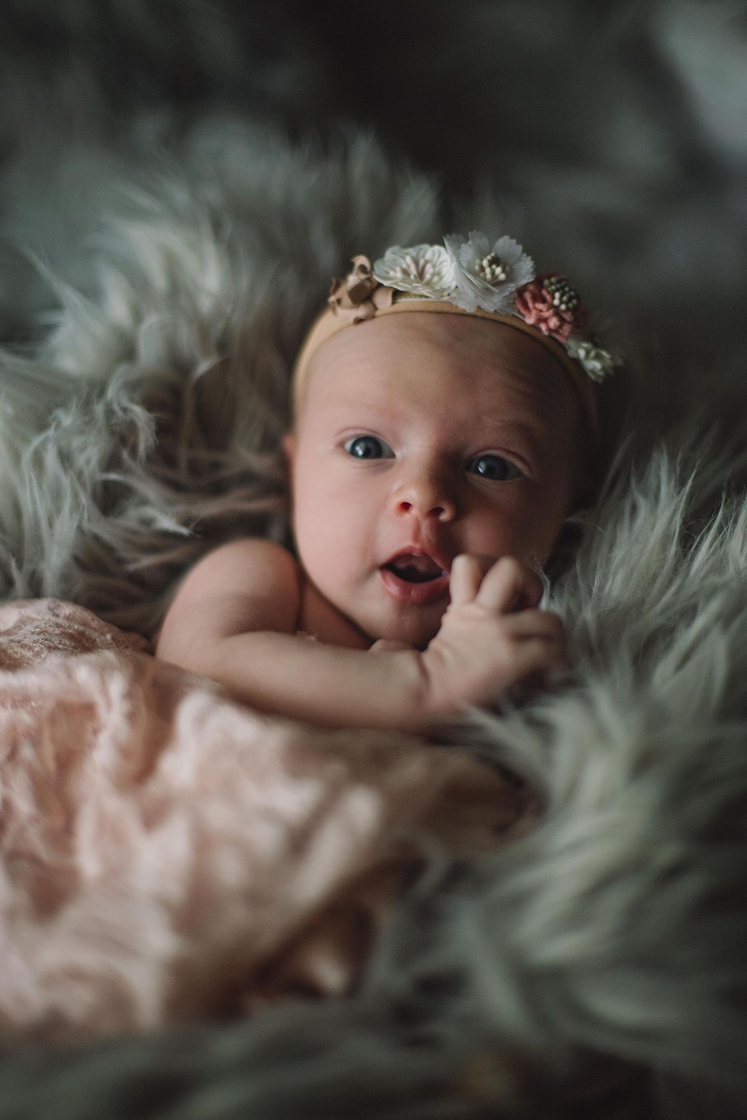 Newborn baby lying on soft grey fur with pink floral headband in dreamy portrait.