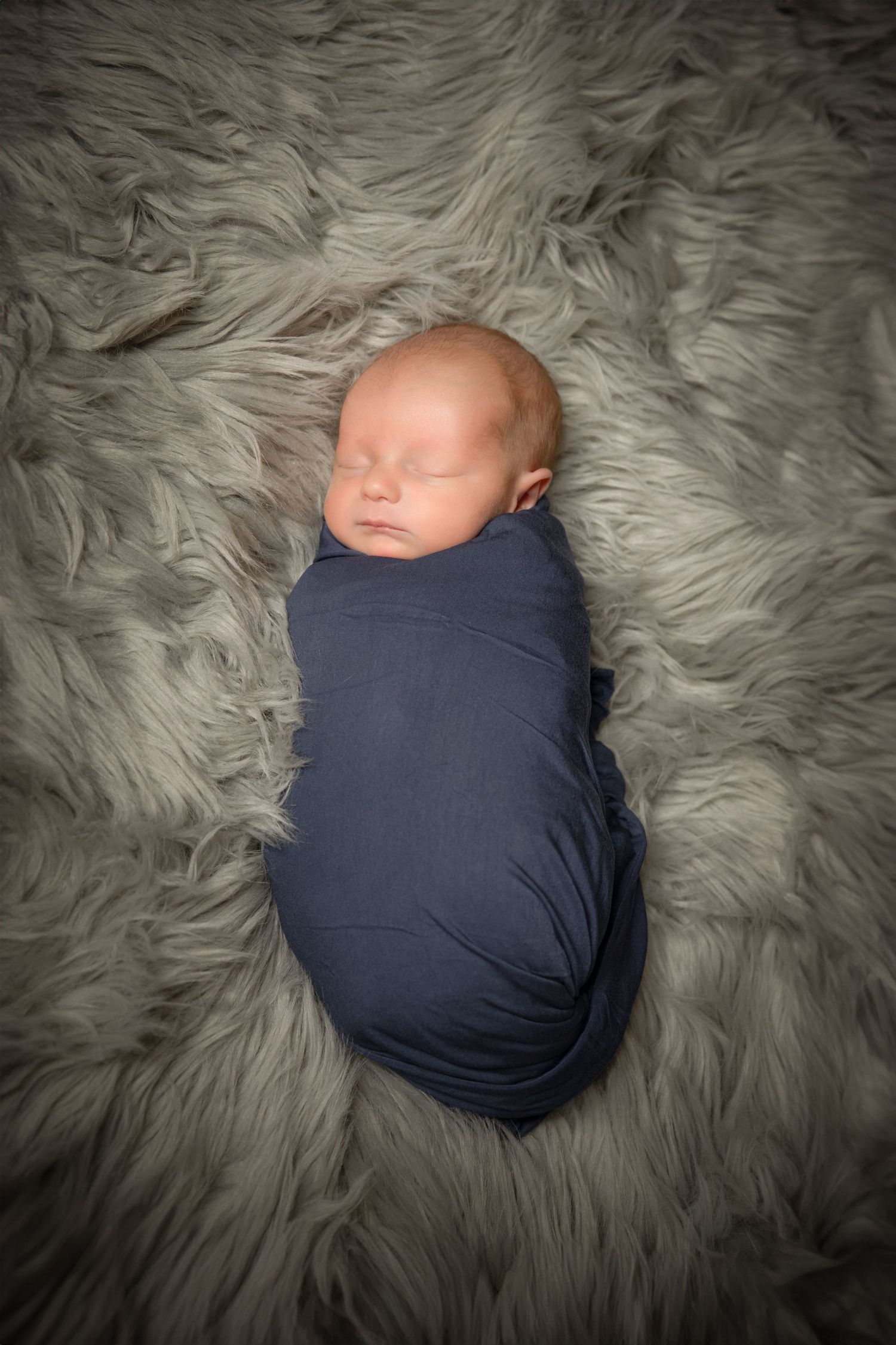 Sleeping newborn baby wrapped in navy blue swaddle blanket lying on soft gray fur backdrop.
