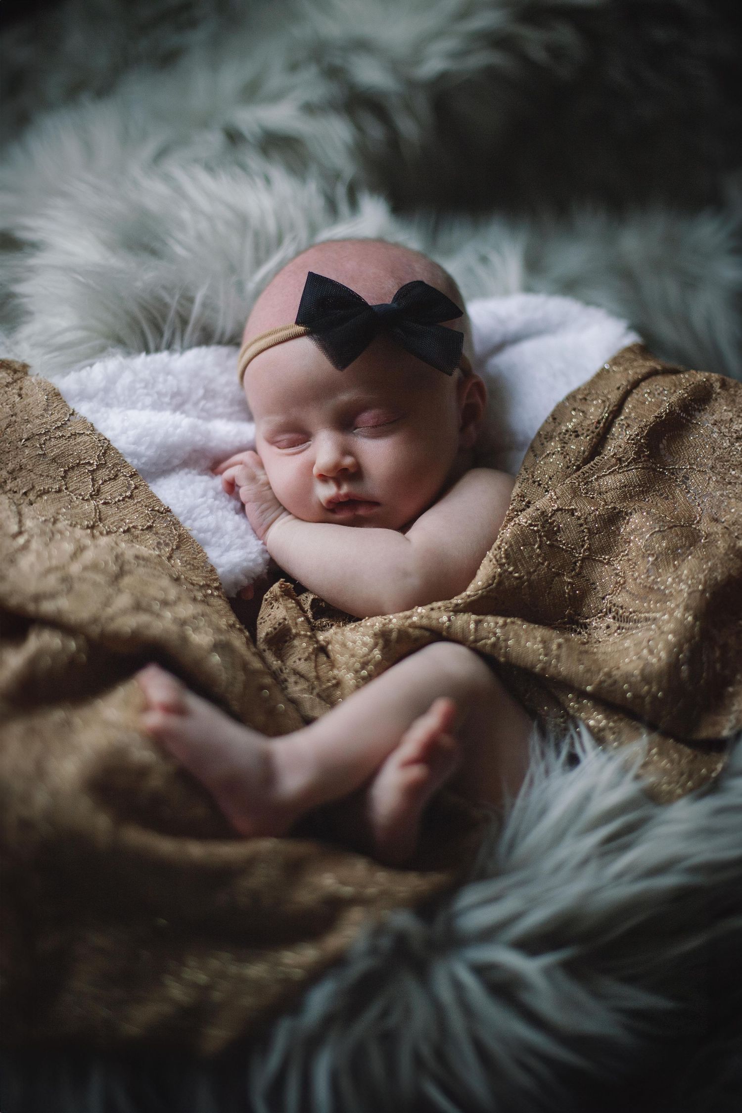 Sleeping baby wearing black bow headband nestles on soft white fur blanket with textured brown backdrop.