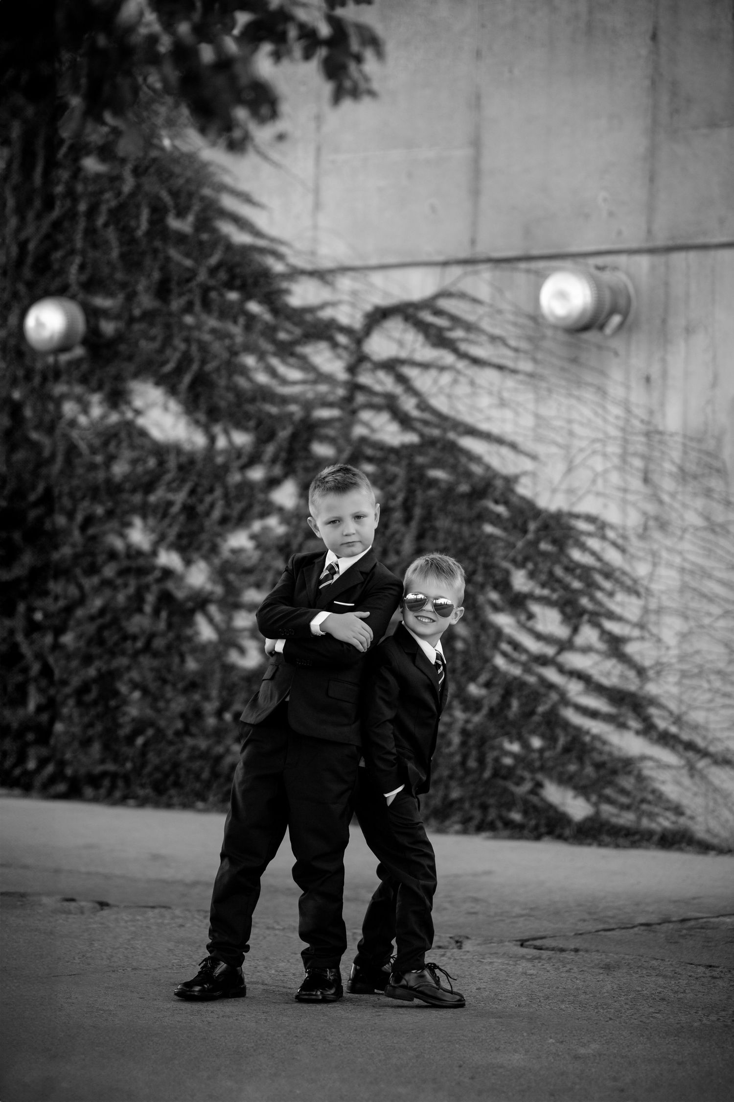 Two young children in suits strike a confident pose together back-to-back against an urban wall at dusk.