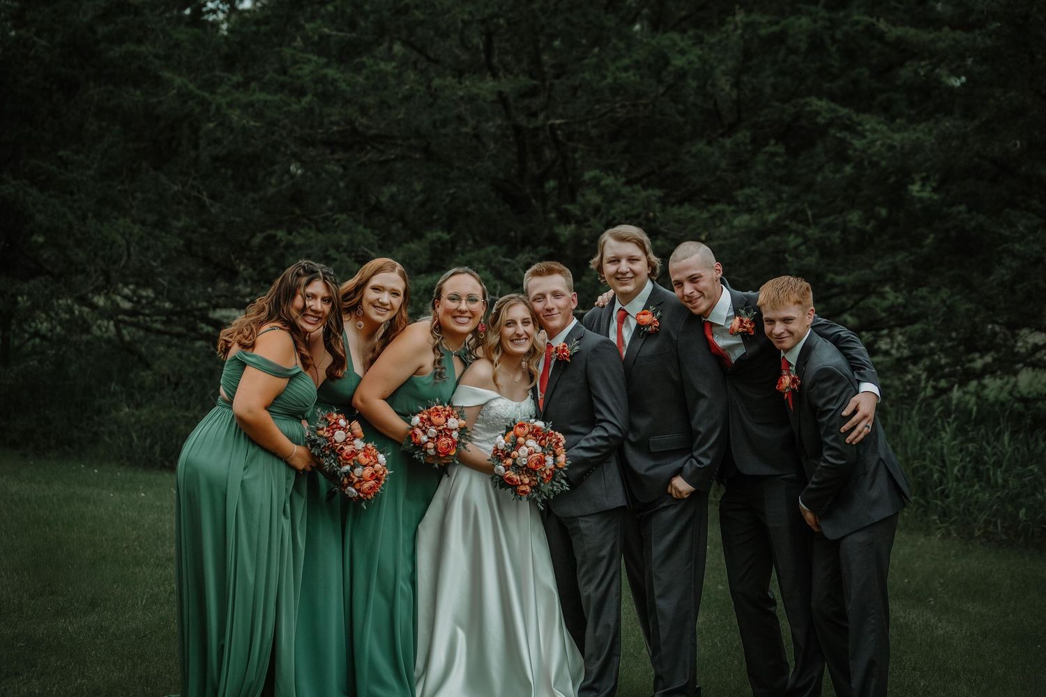 Wedding party in green bridesmaid dresses and dark suits posed together outdoors.