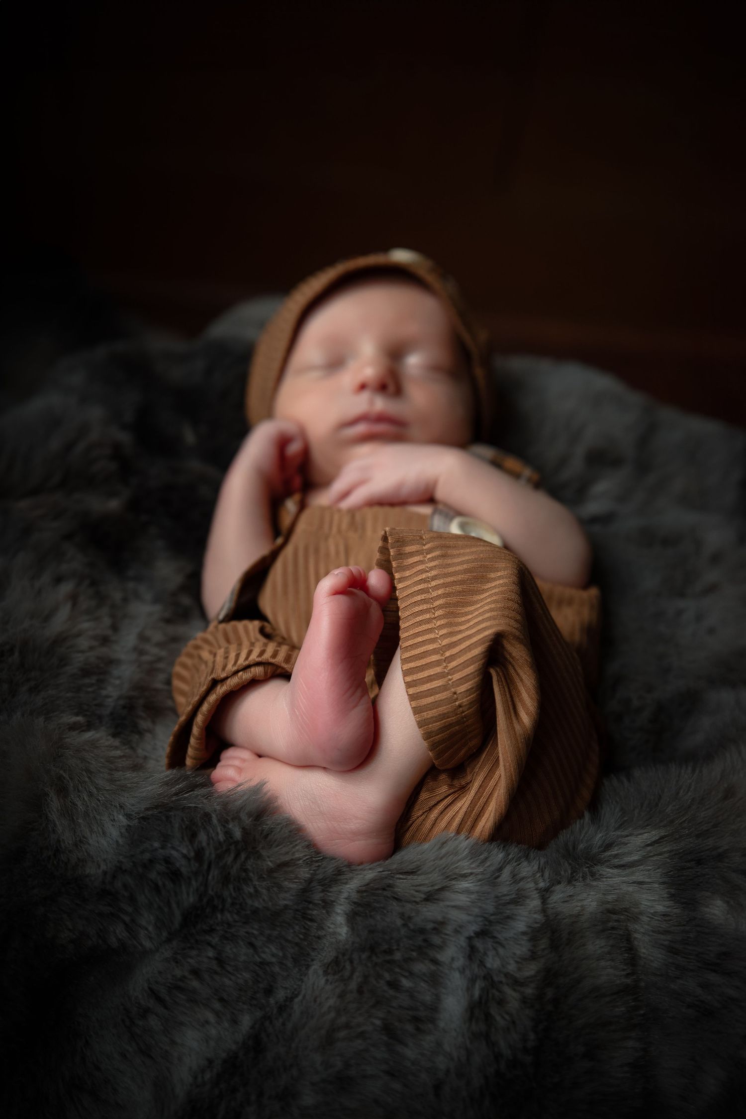 A sleeping newborn baby lies on a dark furry blanket wearing a brown knit sweater in a series of artistic photos.