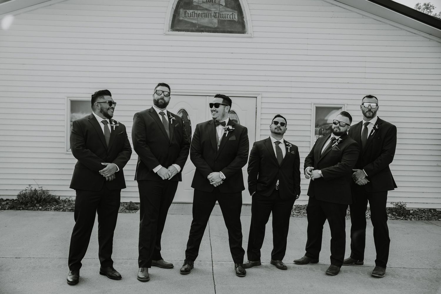 Groomsmen in black suits posing casually outside a white church building.