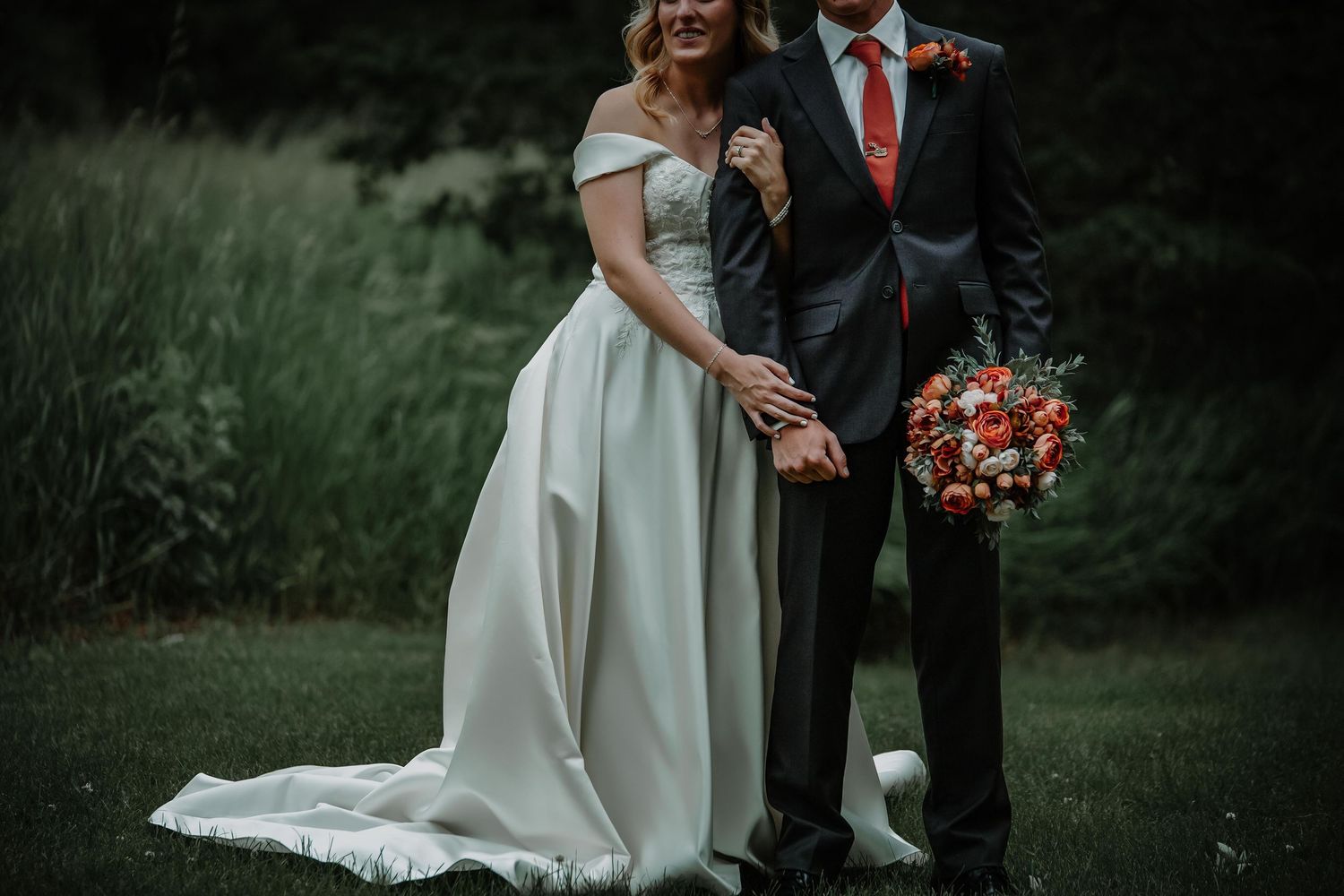 Elegant couple in wedding attire share a moment on grass with coral floral bouquet.