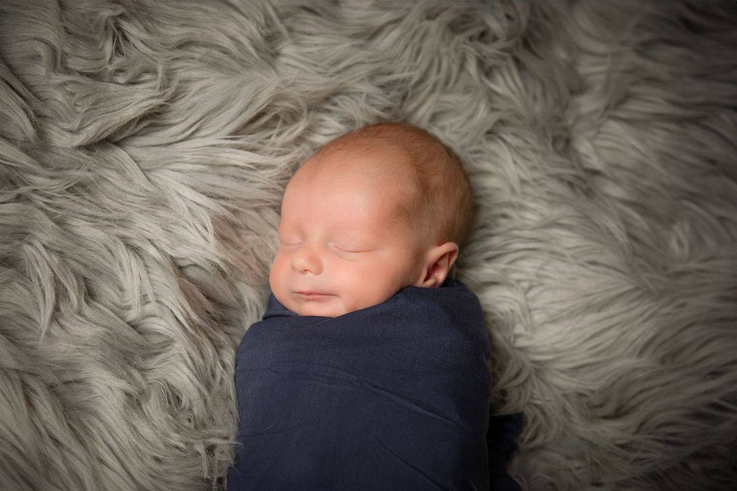 Sleeping newborn baby wrapped in navy blue swaddle blanket lying on soft gray fur background.