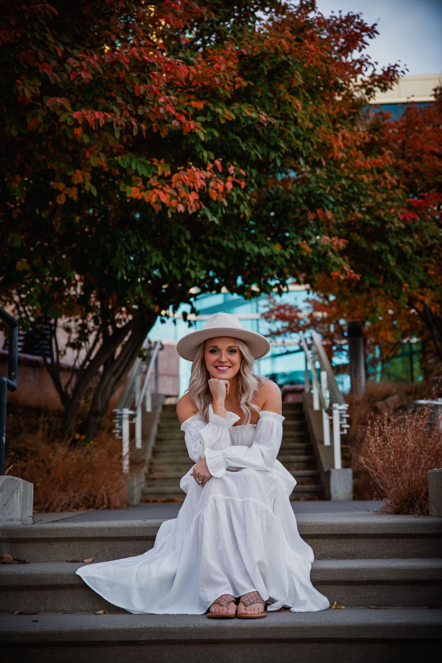 A person in a white dress and hat sits on stone steps surrounded by autumn trees with red and orange foliage.