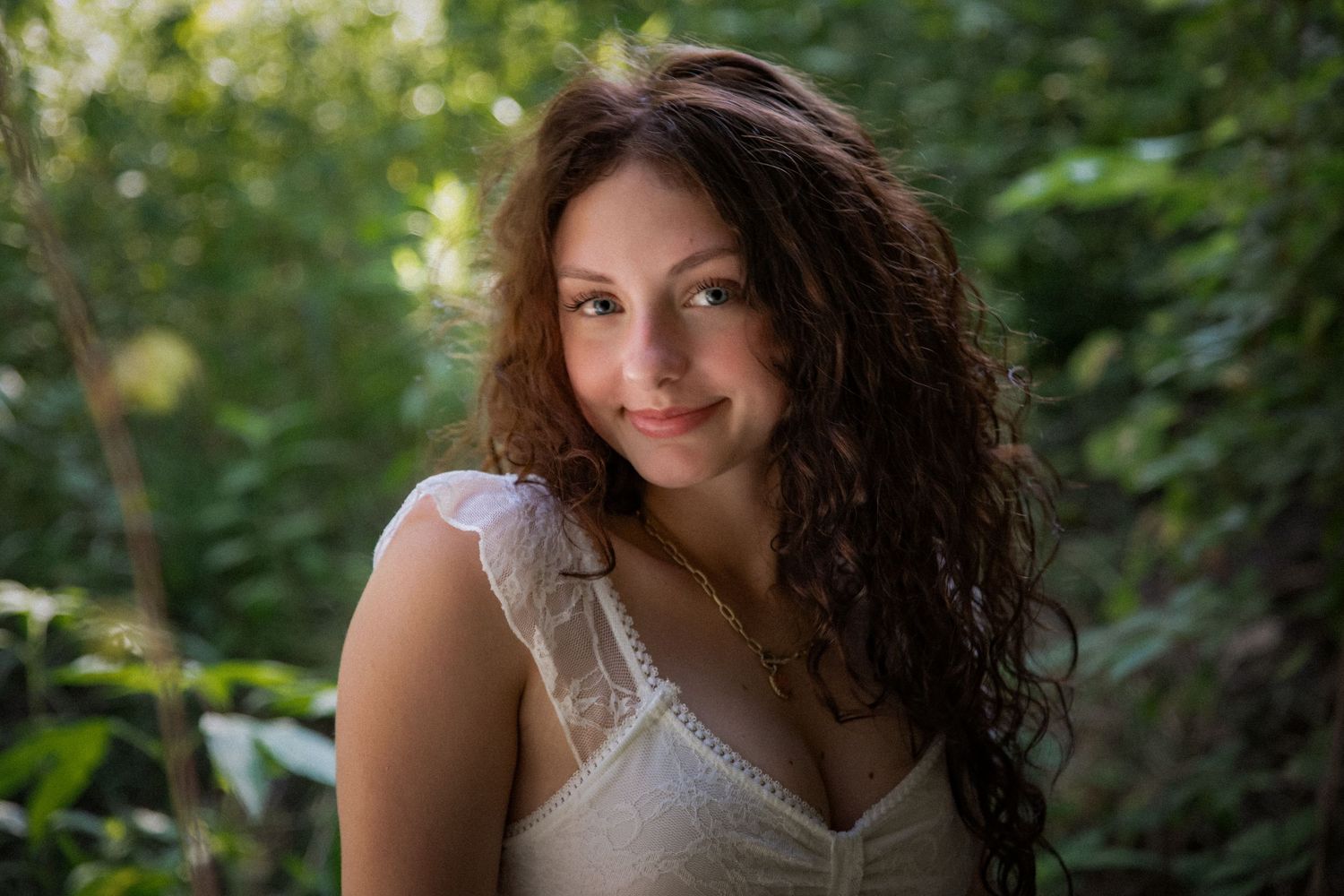Young woman with curly brown hair smiling outdoors, wearing a white lace top surrounded by lush green foliage.