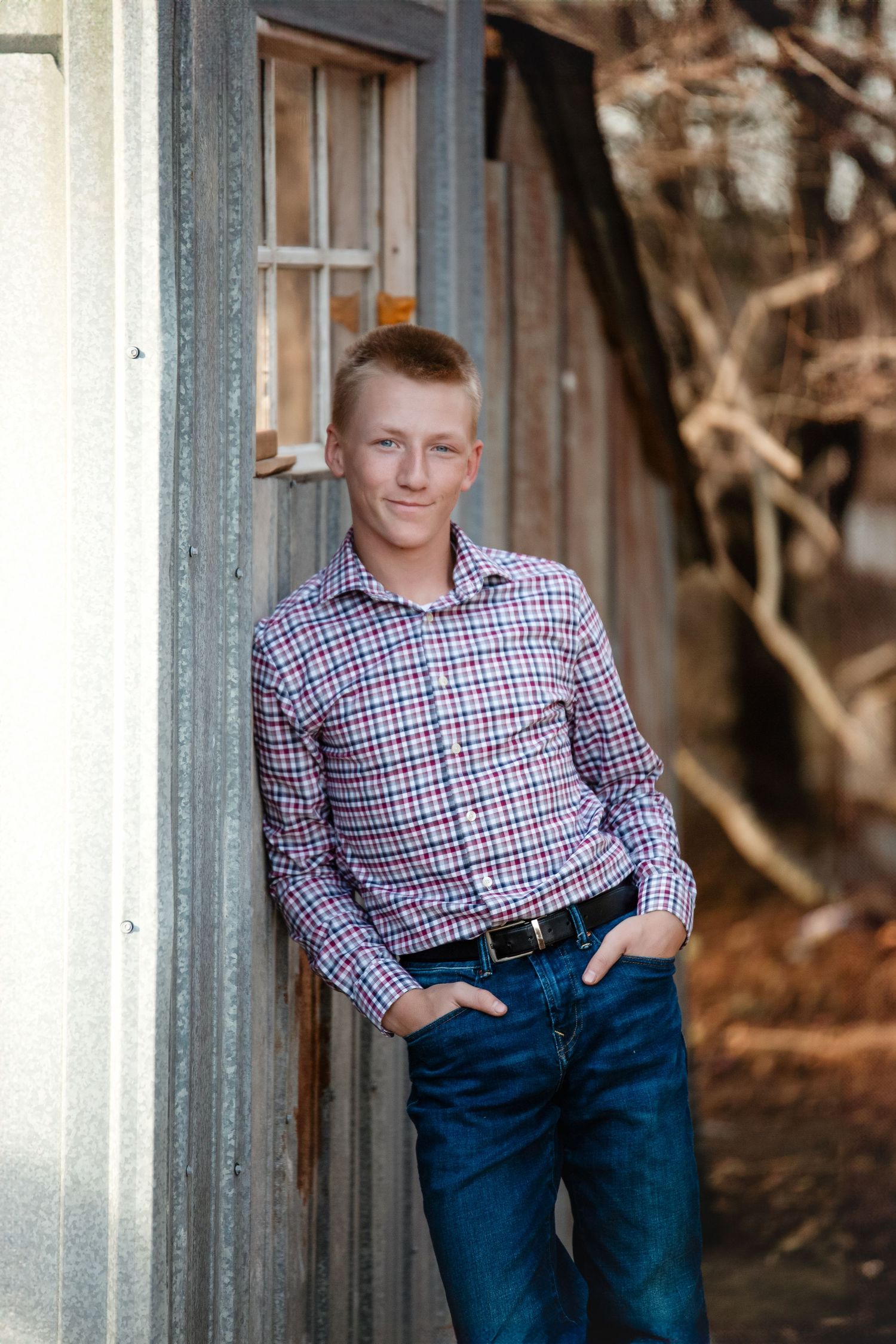 Casual portrait taken outdoors showing person in red and blue checkered shirt with jeans leaning against rustic wooden wall.