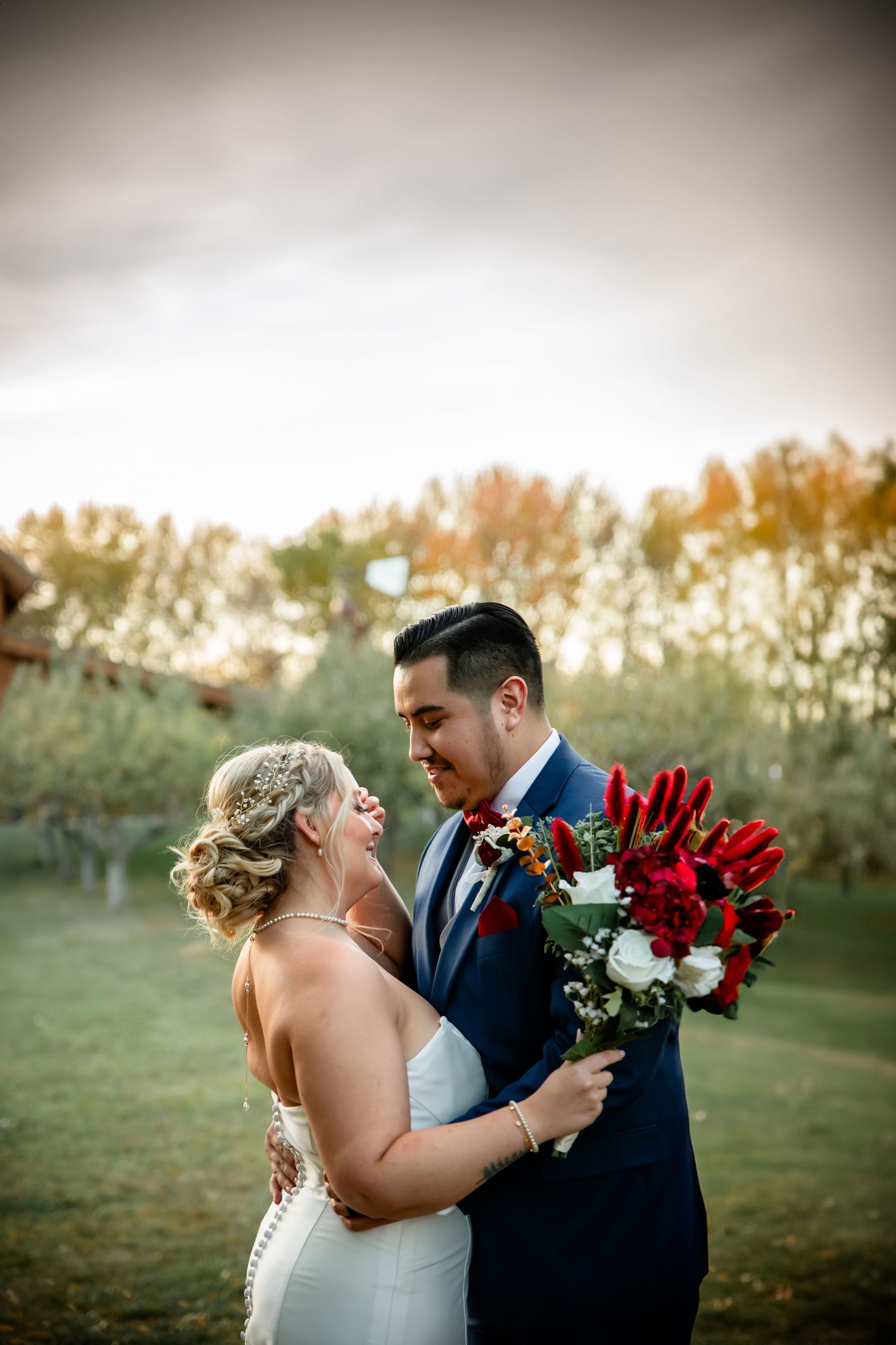 Romantic wedding couple embraces in golden sunset light with red and white rose bouquet.
