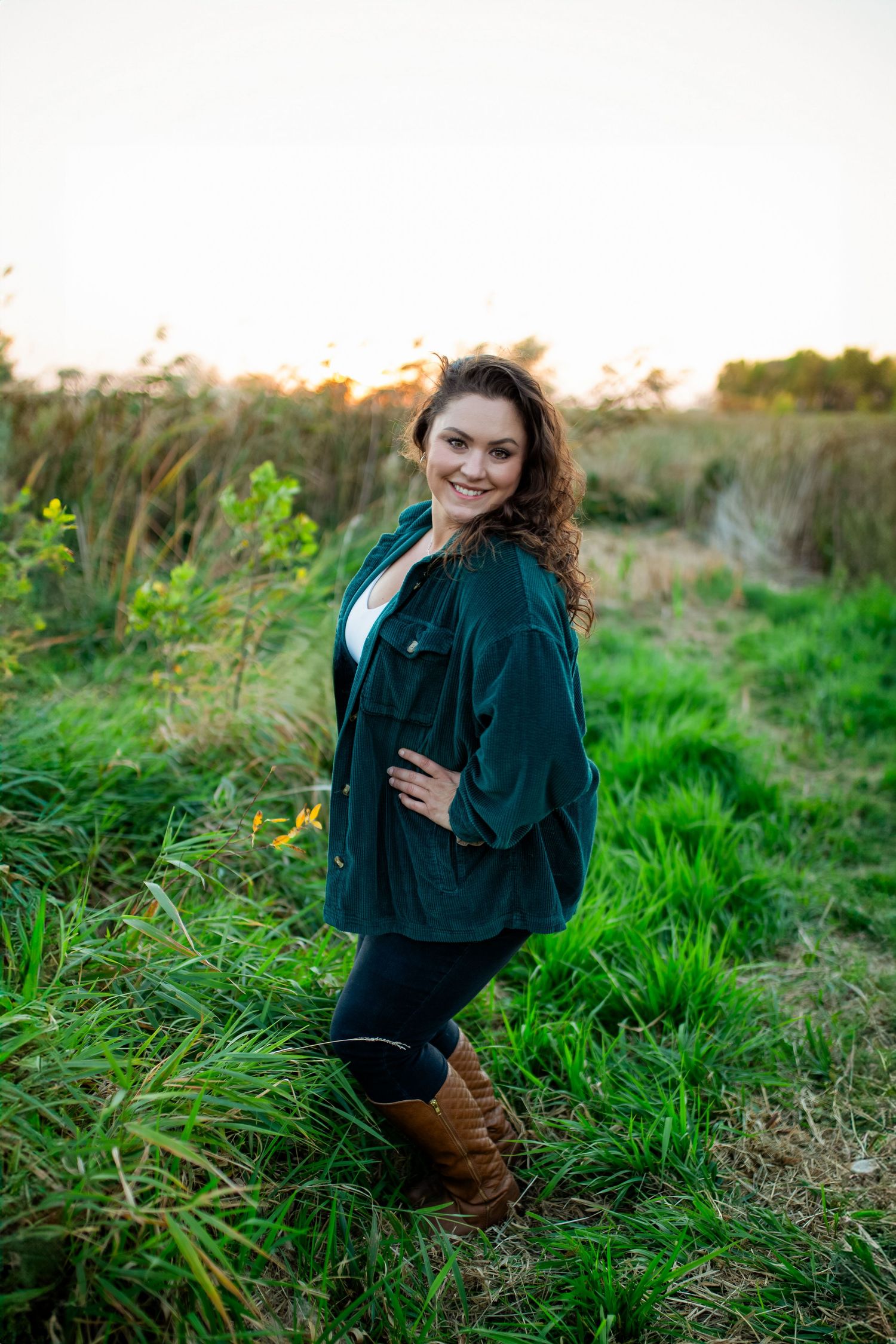Someone poses outdoors wearing a green velvet jacket and brown boots among tall grass at sunset.