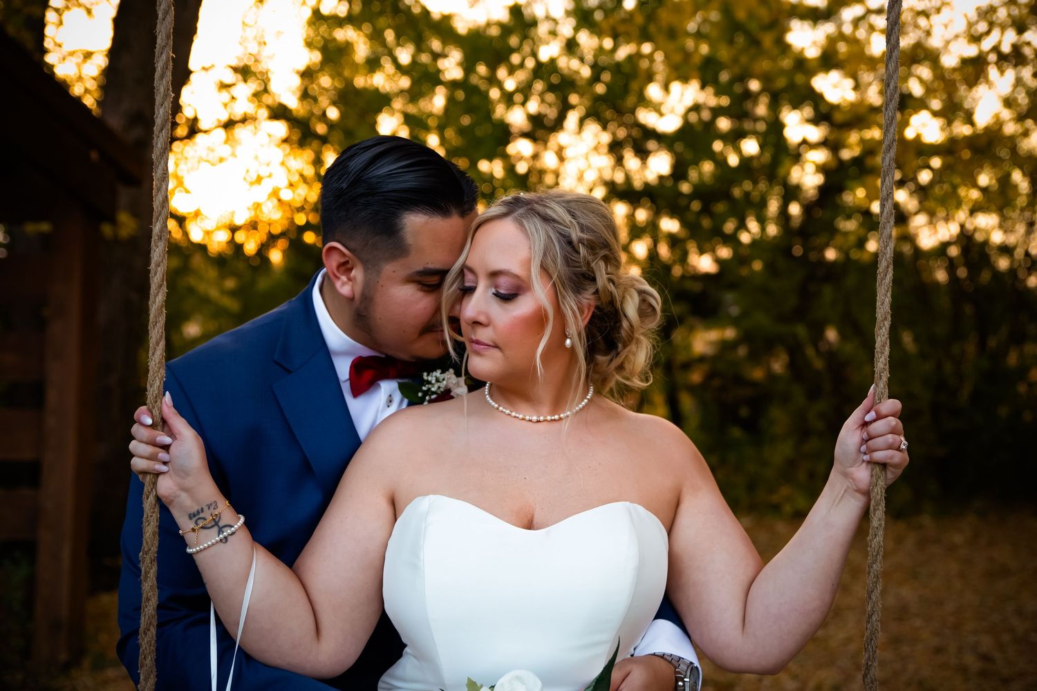 A couple in wedding attire shares an intimate moment during golden hour beneath trees at an outdoor venue.