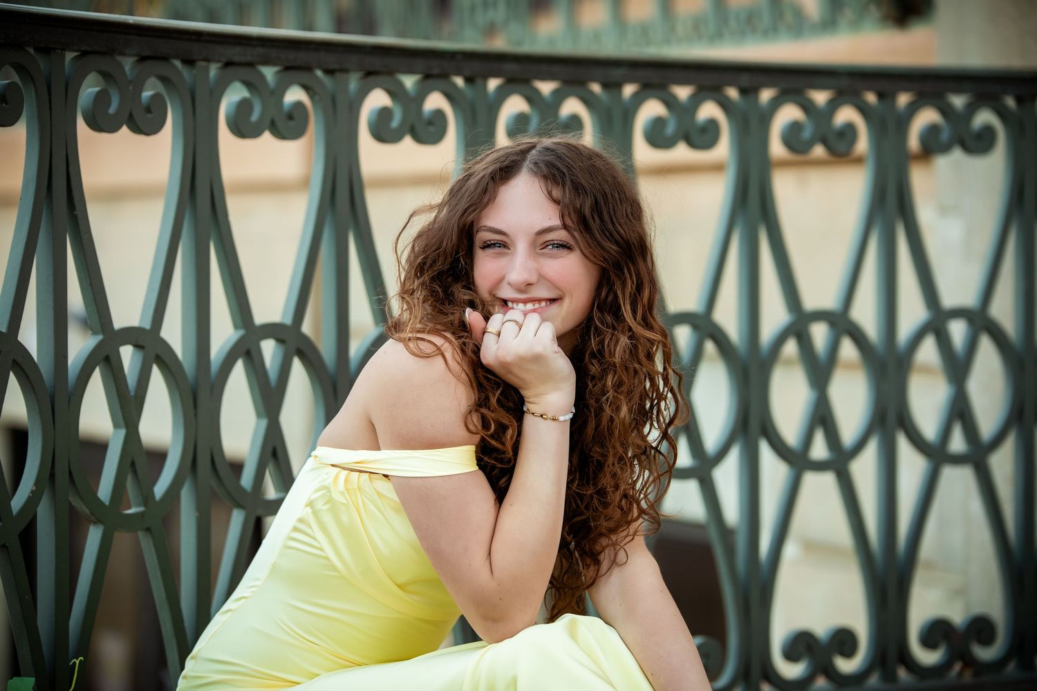 Someone with curly brown hair wearing a yellow dress smiles while sitting near an ornate metal railing.