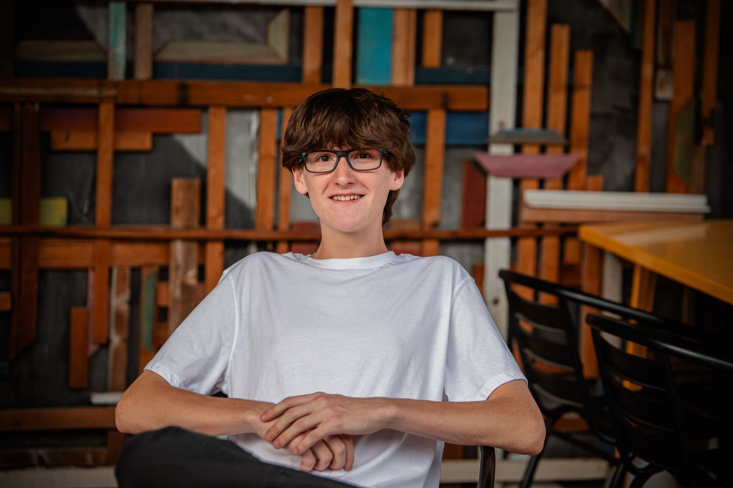 Student wearing glasses sits at wooden desk in library study area with natural lighting coming through windows.